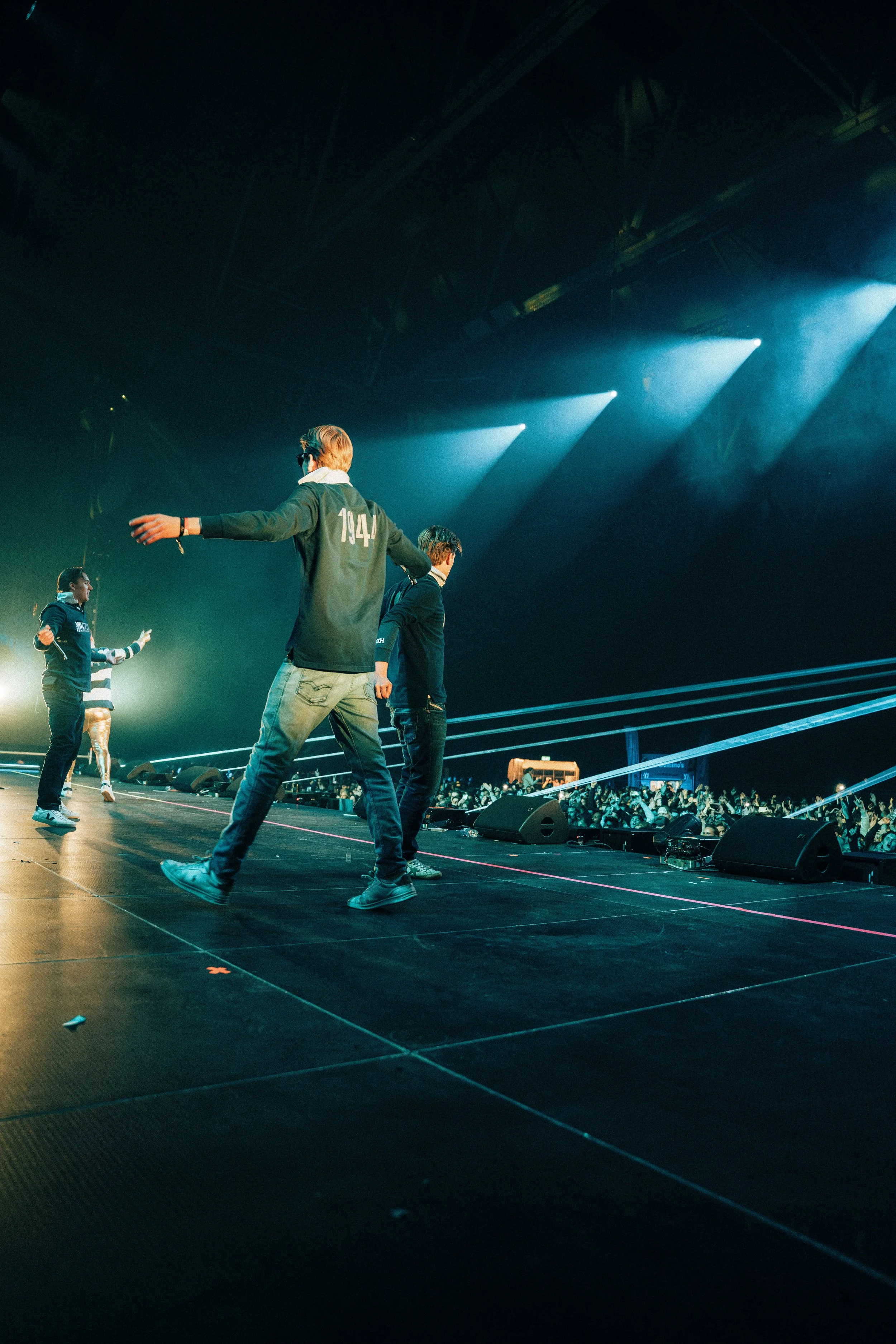 Personer på en scene under konsert, noen danser eller beveger seg til musikk, med lys og publikum i bakgrunnen.