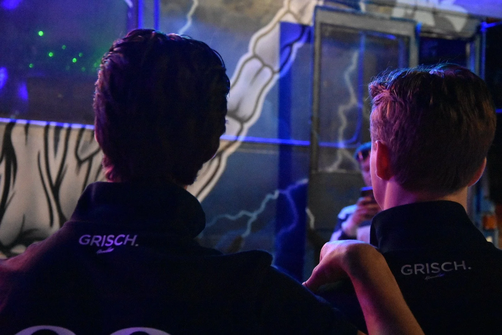 To personer med mørke klær står nær hverandre i en mørk, farget belysning. De har på skuldermerker med teksten 'GRISCH.'