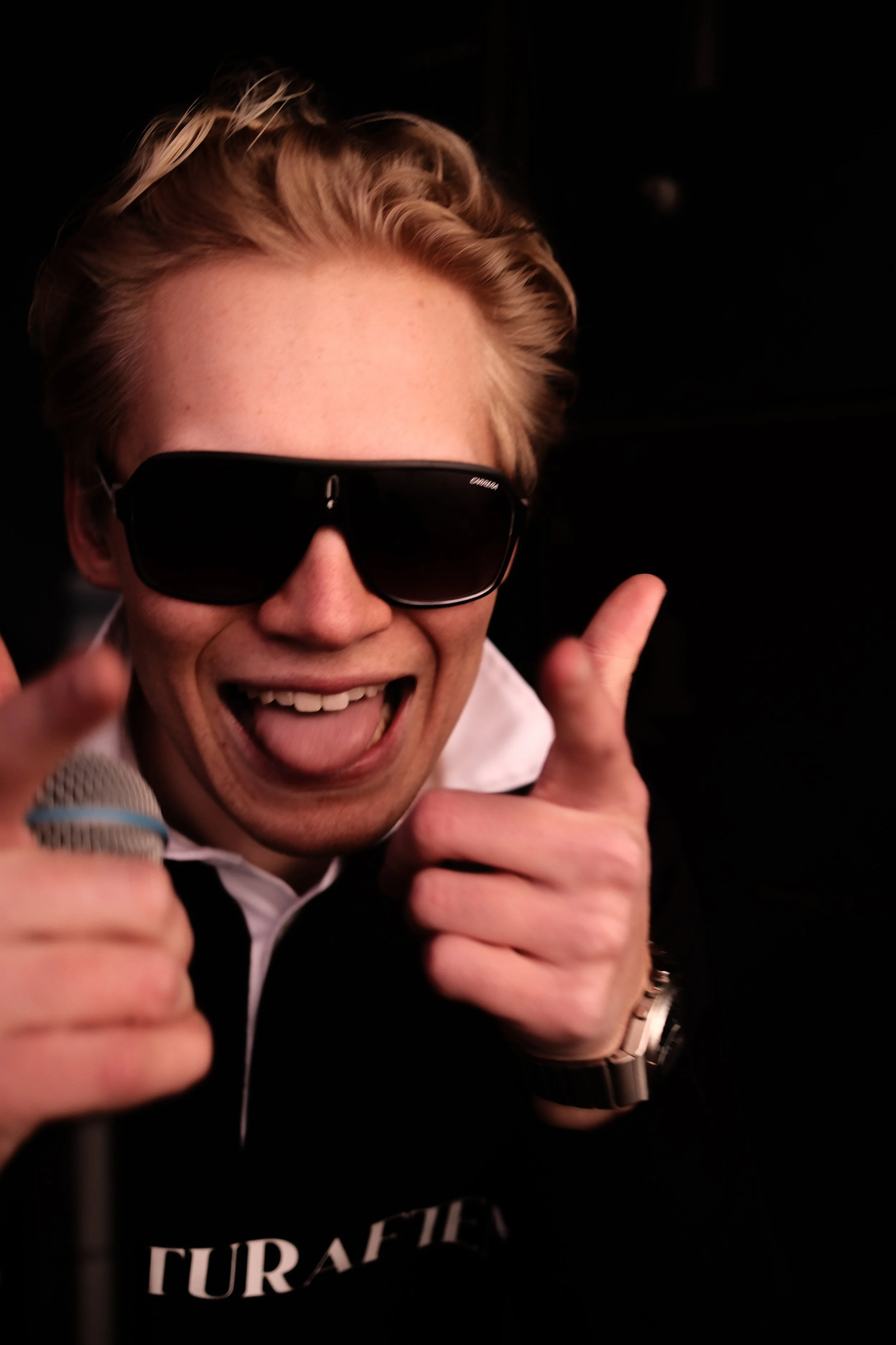 Person med blond hår og svart solbriller som smiler og peker mot kamera.