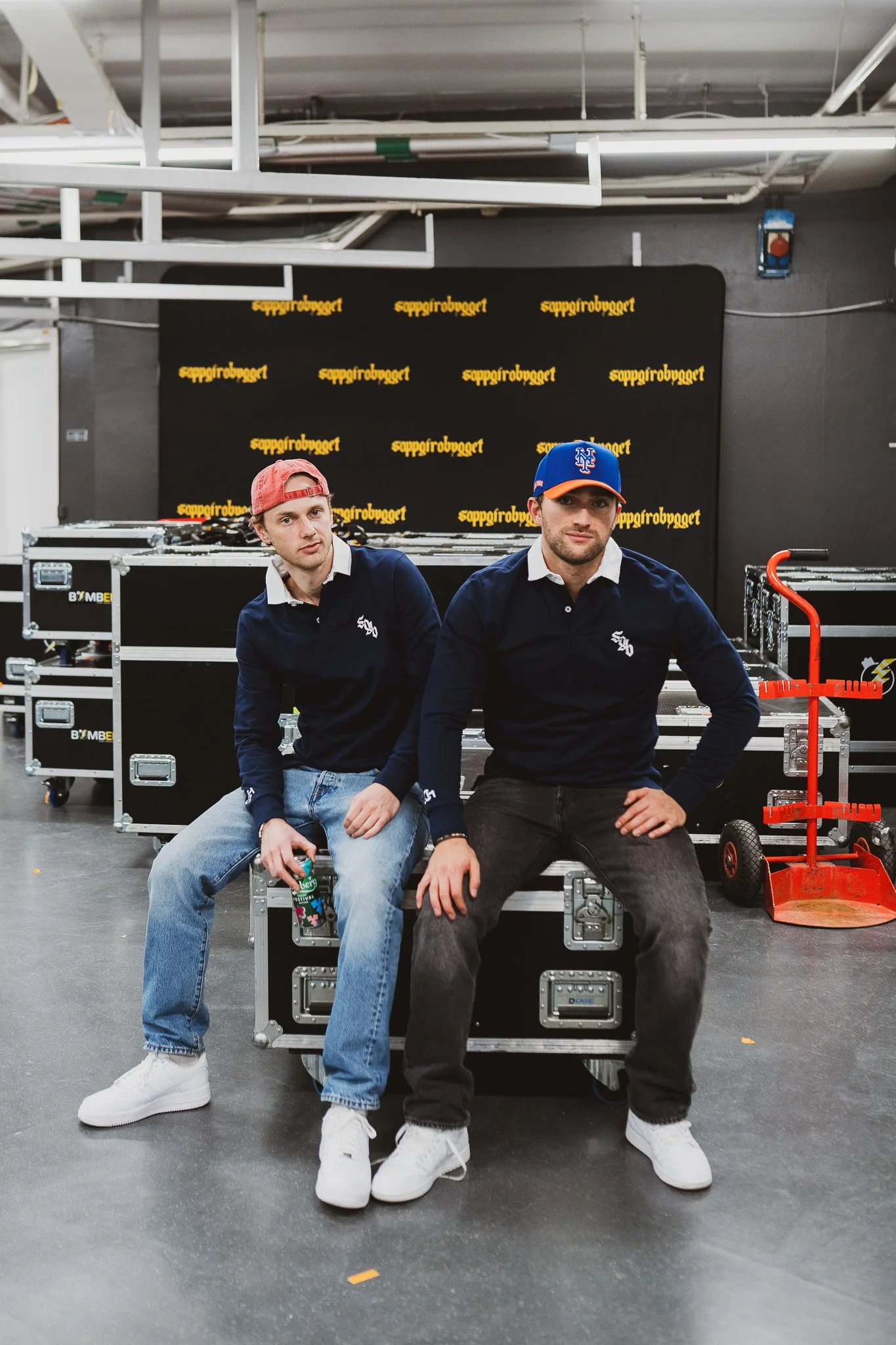 To unge menn sitter på en metallkasse i et rom med produksjonsutstyr og et svart bakteppe medLogo. Begge har på seg mørke gensere og hvite sneakers. Mannen til venstre har hvit skjorte under gensaren, rød caps, og holder en vannflaske. Mannen til høgre har blå caps med oransje visir og ser direkte på kamera.