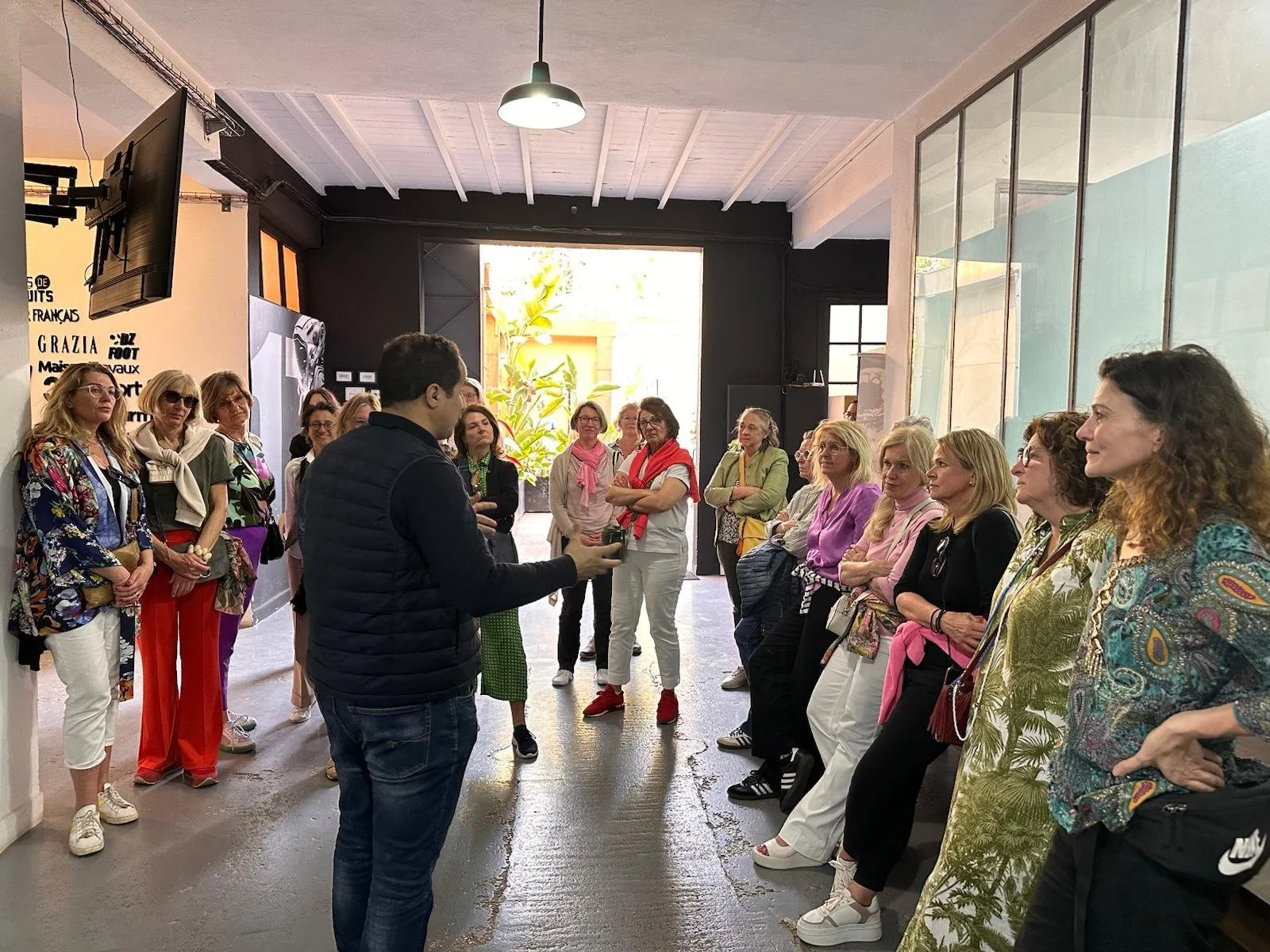 Eine Gruppe von Frauen hört aufmerksam einem Mann zu, der eine Präsentation hält, in einem modernen Raum mit großen Fenstern und plakatenartigen Wanddekorationen.