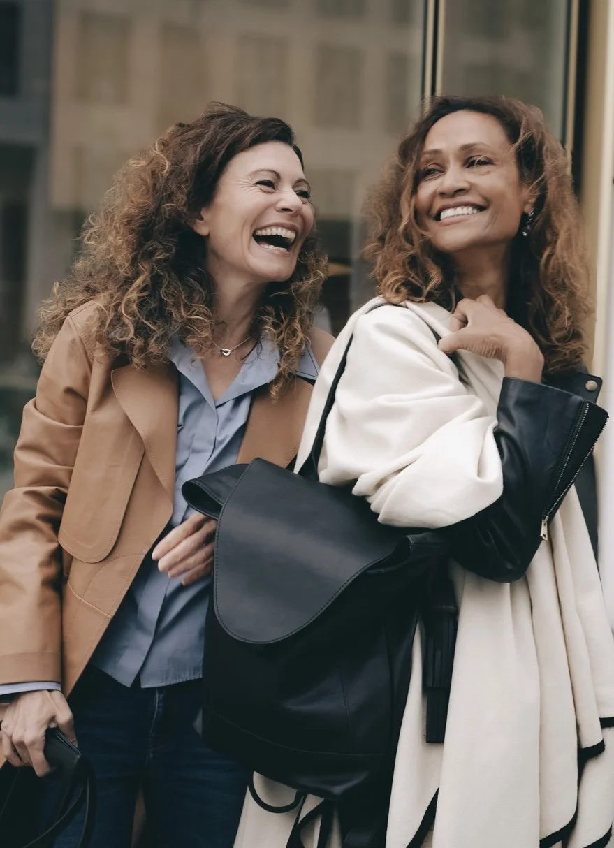 Zwei Frauen, eine weiss mit lockigen Haaren und braunem Mantel, eine mit brauner Haut lachen gemeinsam.