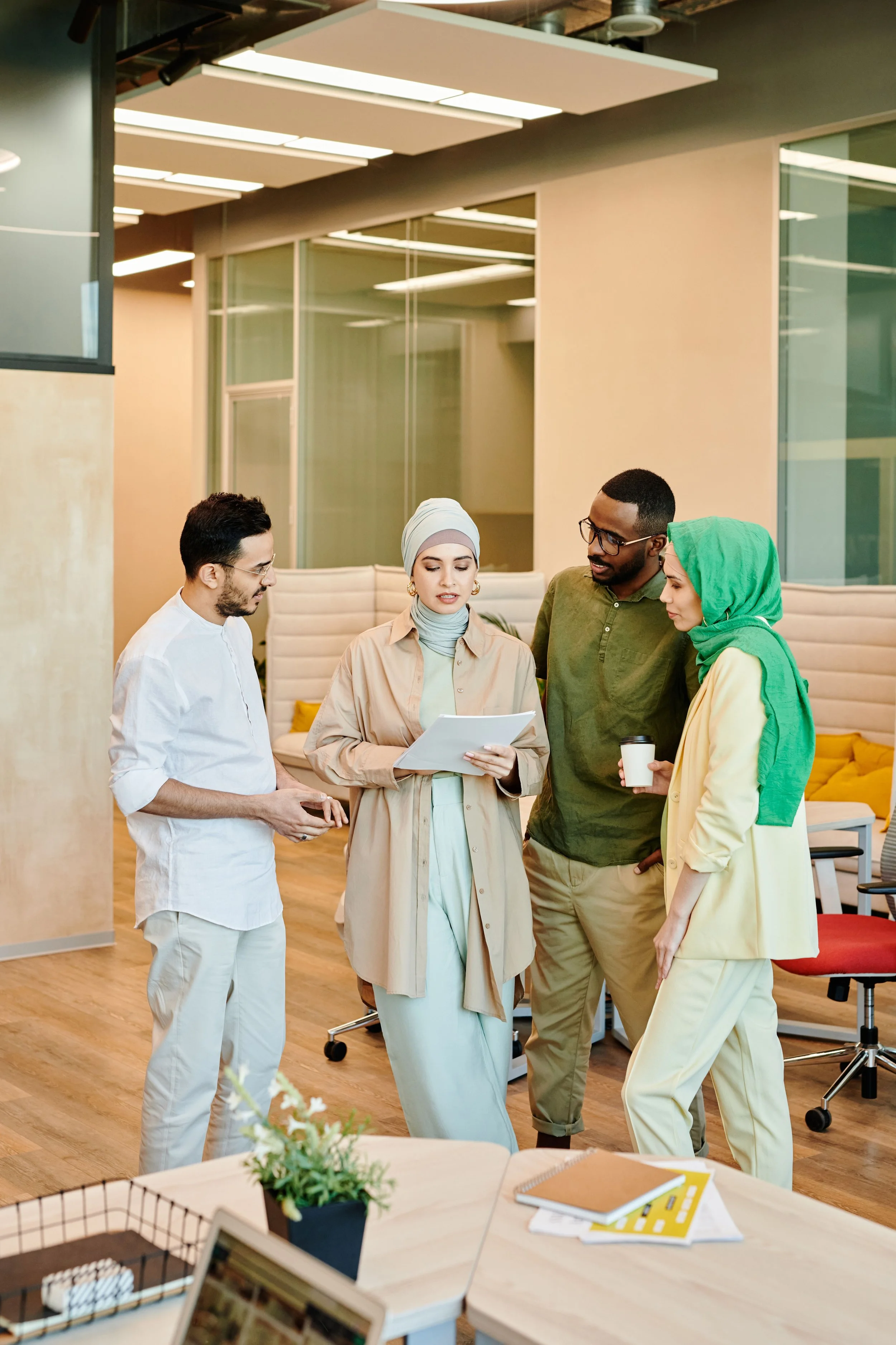 Zwei Frauen mit Hijab stehen zusammen mit zwei Männern in einem Büro und besprechen ein Papier, das eine der Frauen in der Hand hält.