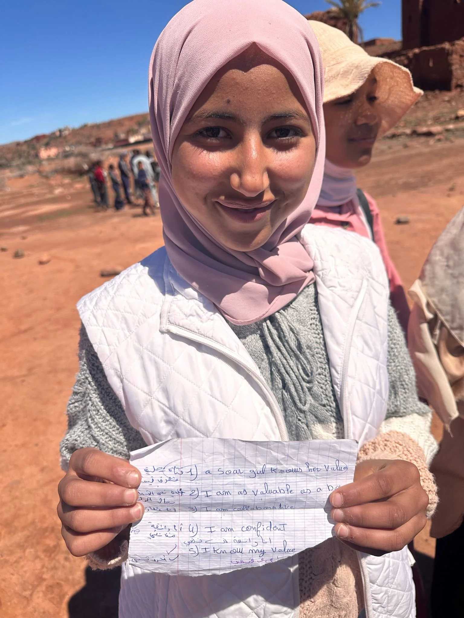 Ein Mädchen trägt einen hellrosa Schal und hält ein handgeschriebenes Blatt Papier mit englischem Text. Im Hintergrund sind andere Personen in einer Westsahara-Wüstenlandschaft bei sonnigem Wetter zu sehen.