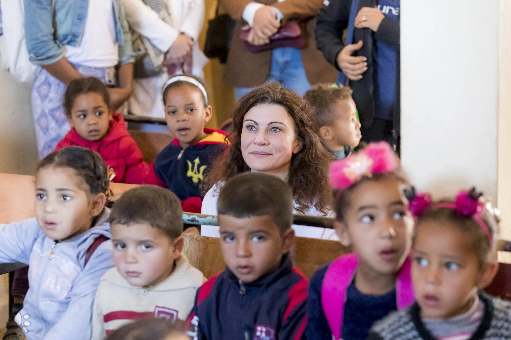 Eine Gruppe von Kindern sitzt in einem Raum mit einer erwachsenen Frau, die lächelt, während die Kinder neugierig und aufmerksam schauen.