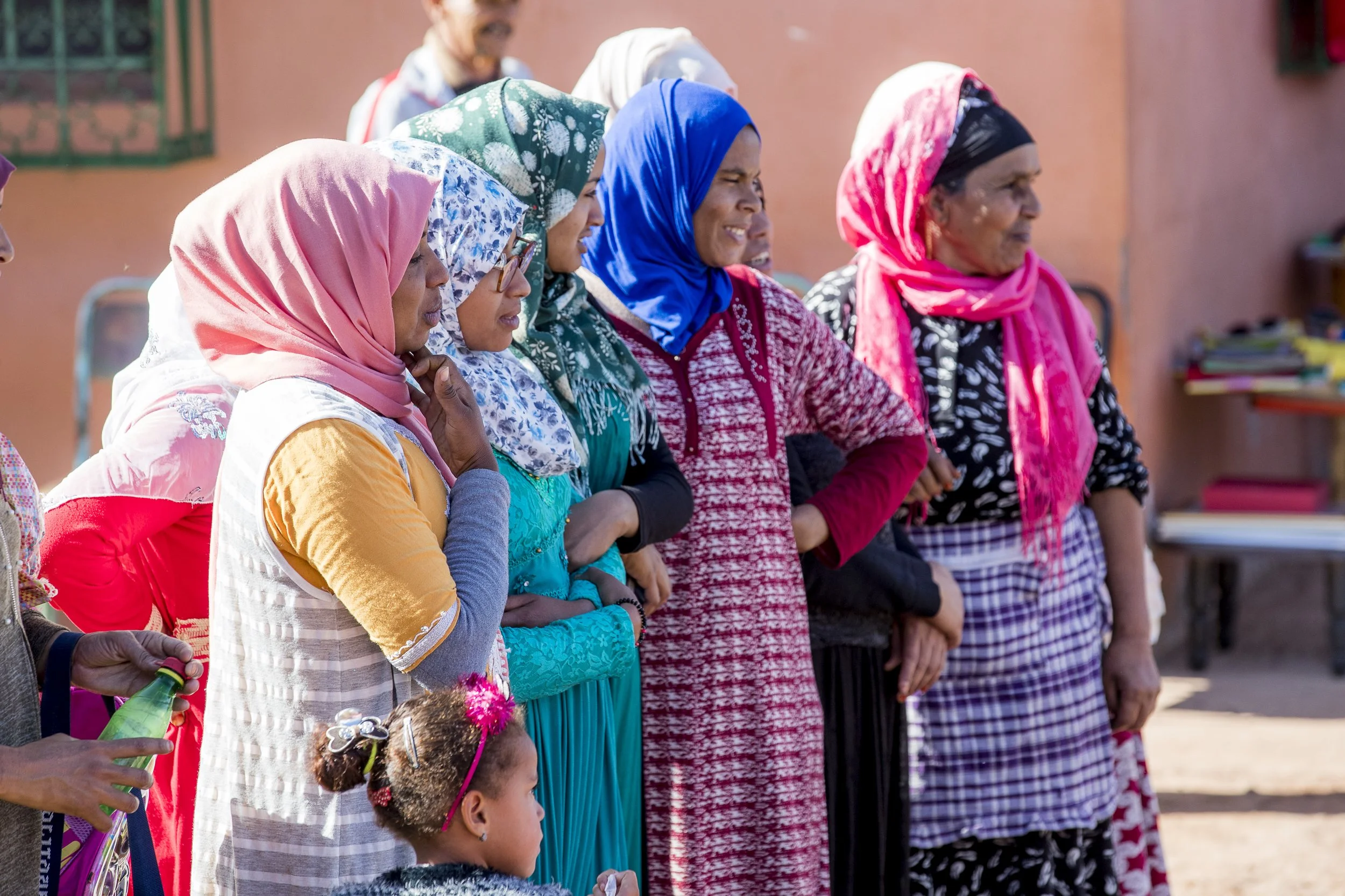 Mehrere Frauen und Kinder stehen in einer Reihe und schauen aufmerksam nach rechts. Sie tragen bunte Kopftücher und Kleidung, im Hintergrund eine sandfarbene Wand.