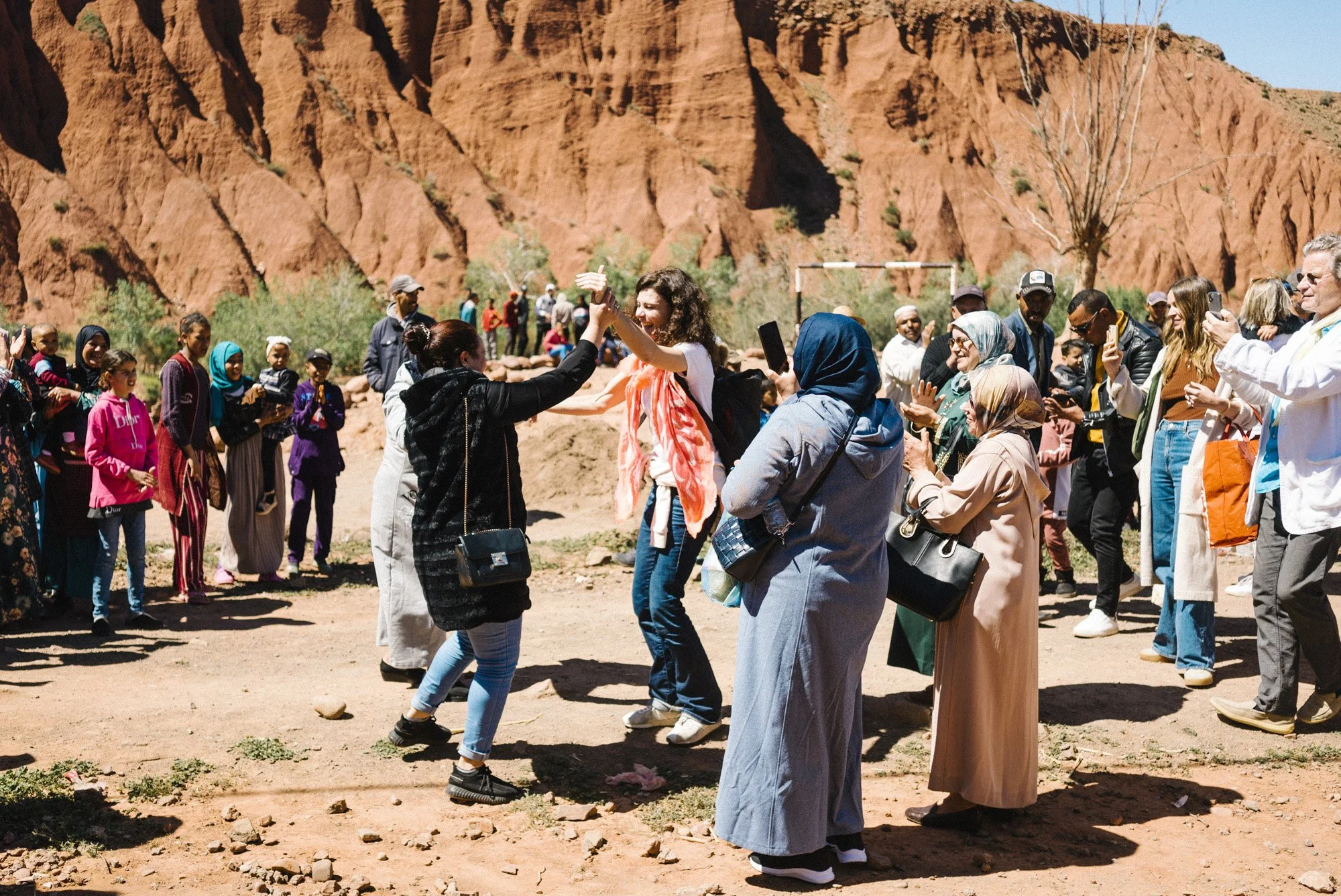 Gruppe von Menschen, die sich in einer Wüstenlandschaft mit roten Felsen versammeln, einige begrüßen sich, andere fotografieren, im Hintergrund hebt sich eine kahle Birke vor der felsigen Landschaft ab.