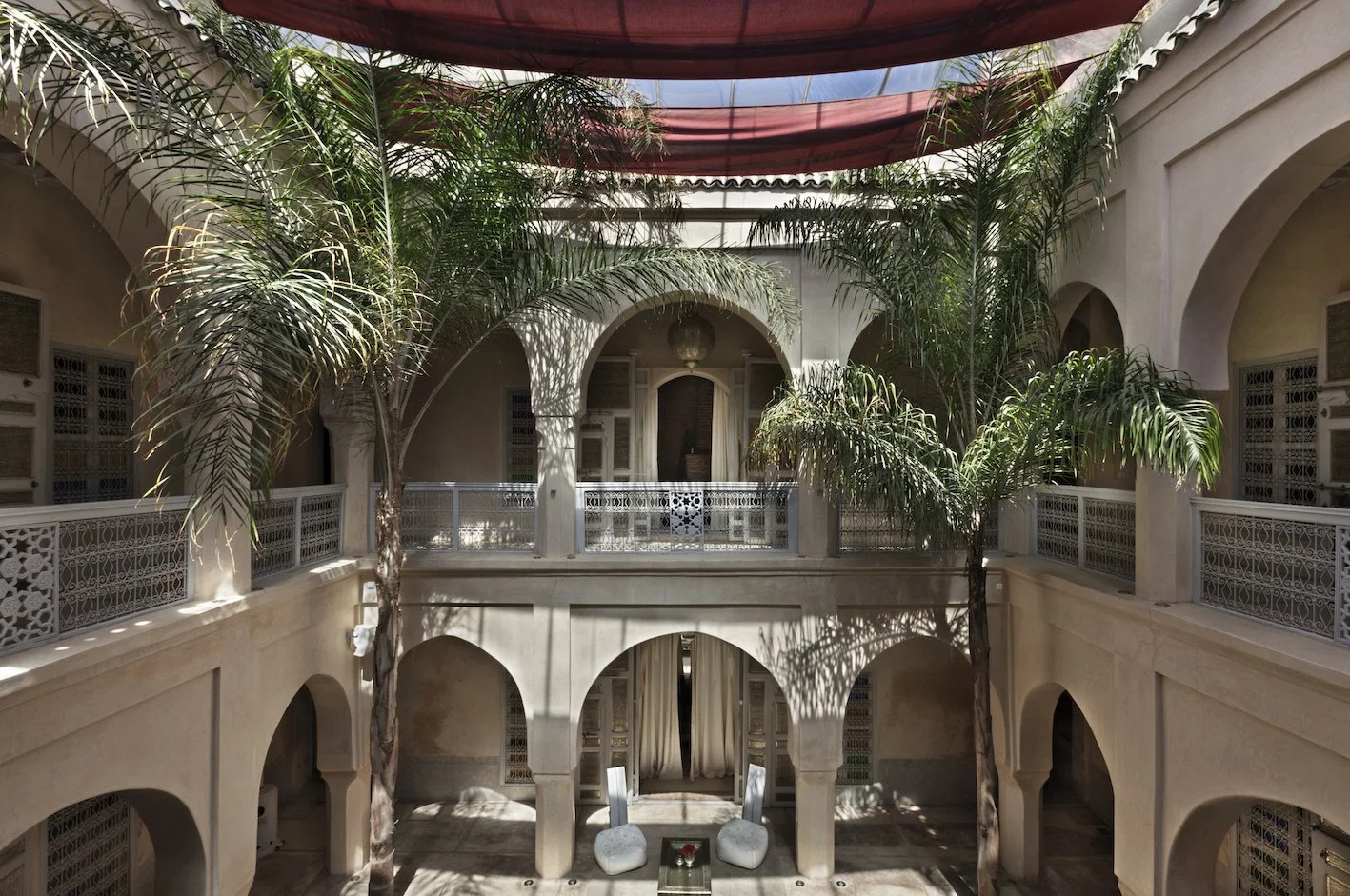Innenhof eines Riads in Marrakesch. Man sieht zwei Palmen sowie rote Drapées über einem offenen Innenhof. Die Wandfarbe ist creme.
