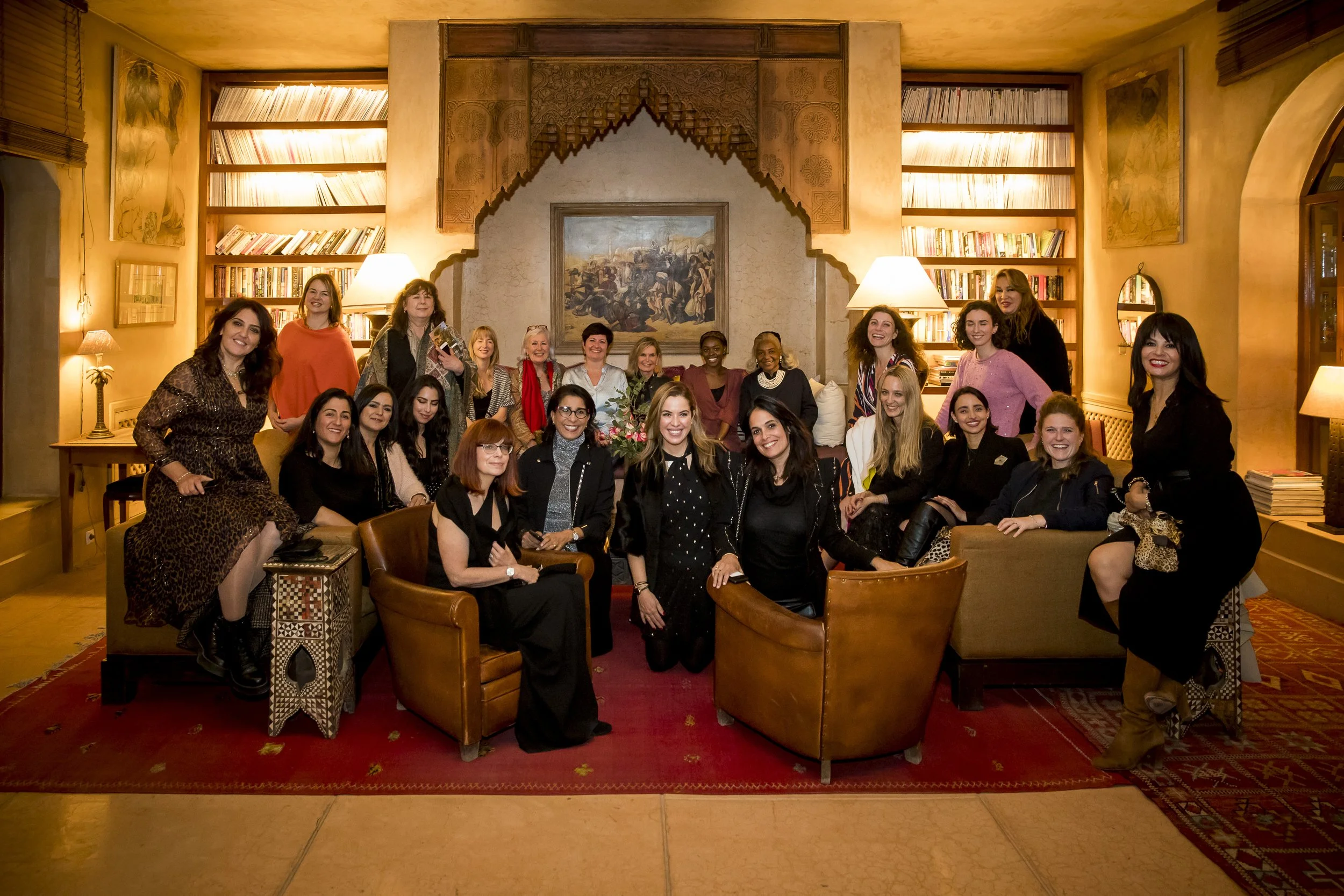 Eine Gruppe von Frauen sitzt und steht in einem gemütlichen, warm erleuchteten Raum mit Büchernregalen, einem Gemälde und Lampen.