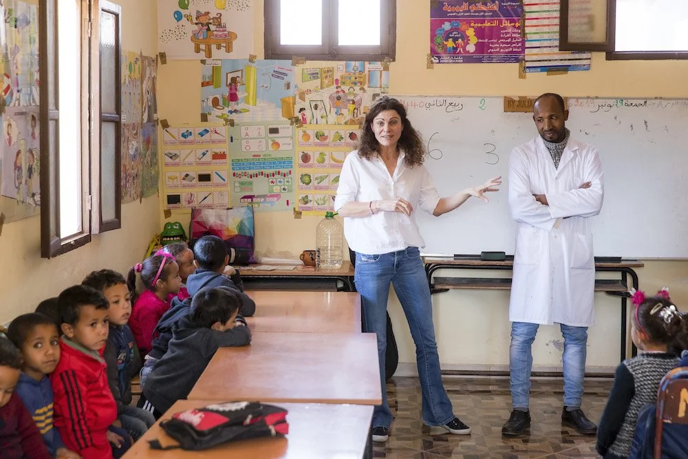 Eine Lehrerin zeigt einer Gruppe von Schulkindern in einem Klassenraum etwas an der Tafel, während ein Lehrer neben ihr steht. Die Kinder sitzen an Tischen und schauen aufmerksam zu. Der Klassenraum ist bunt dekoriert mit Postern und Zeichnungen an den Wänden.