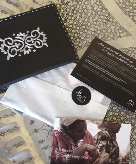 Eine schwarze Tasche mit weißen Dekorationen, eine Karte, eine silberne Geschenkverpackung, gesticktes Stoffmuster, und ein Foto von zwei Menschen, die ein Herz formen.