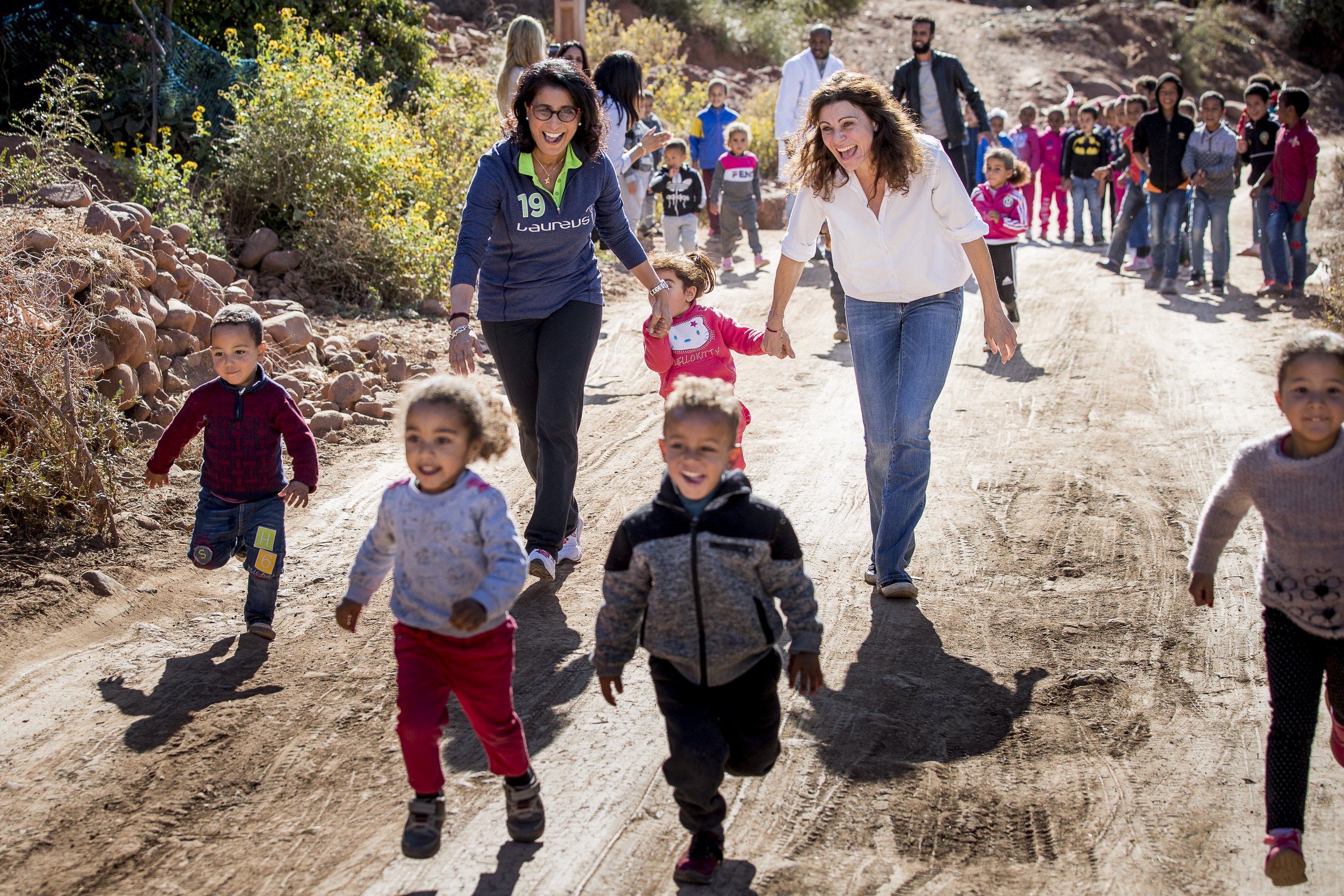 Eine Gruppe von Kindern und Erwachsenen laufen lachend auf einem Sandweg in der Natur, umgeben von Büsche und Steinen, bei sonnigem Wetter.