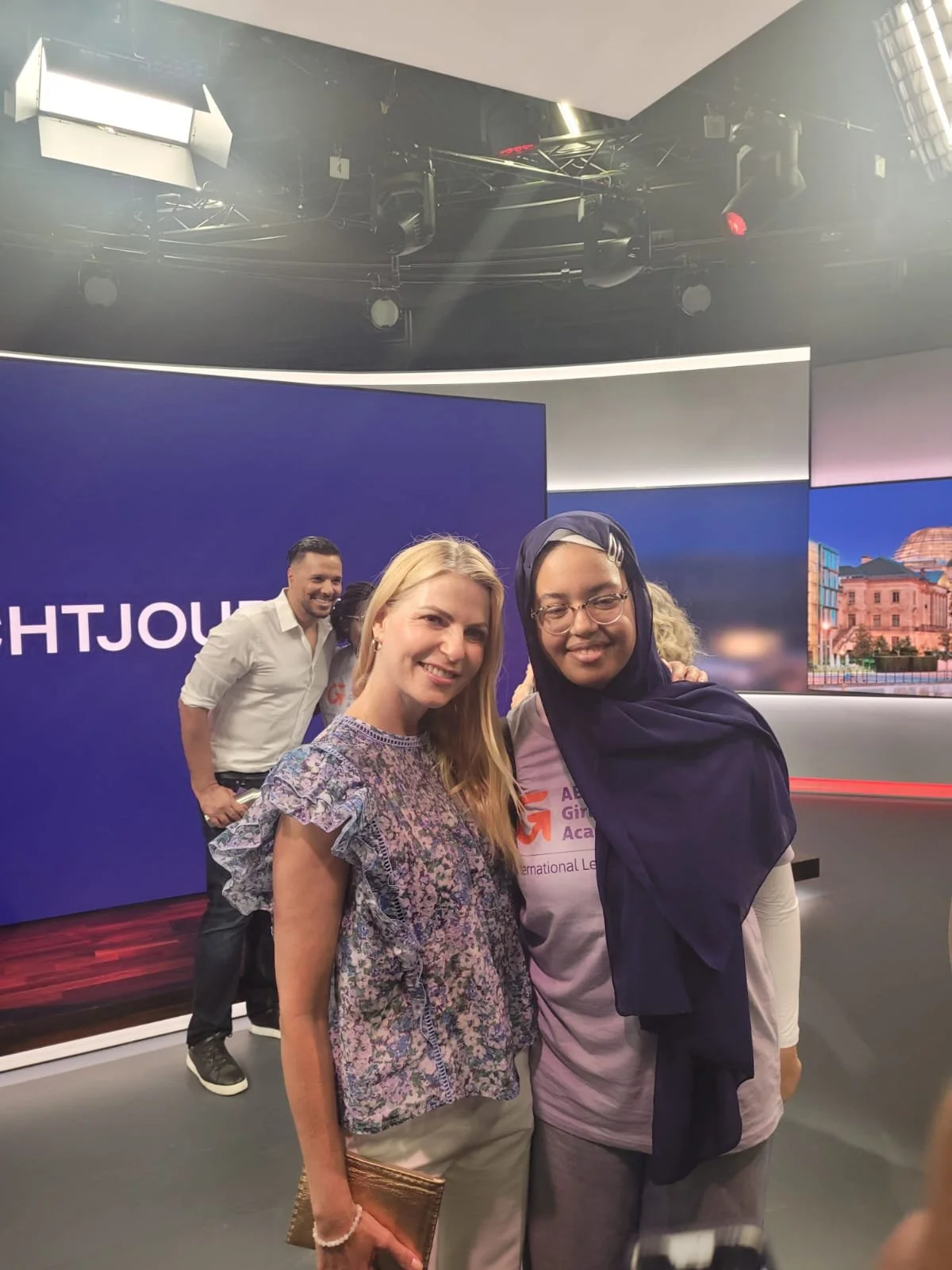 Drei Menschen, zwei Frauen und ein Mann, posieren vor einem blauen Hintergrund bei einem Event. Die Frauen sind lachend, die Frau auf der rechten trägt ein Kopftuch. Im Hintergrund ein Raum mit Kamera- und Lichttechnik.
