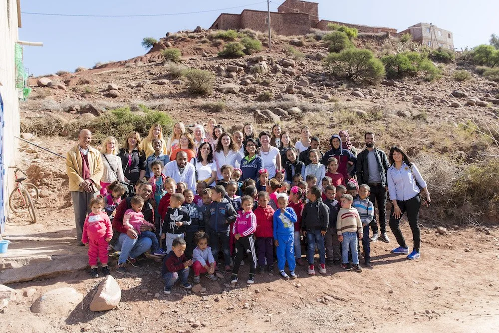 Gruppenfoto von Erwachsenen und Kindern vor einem Hügel mit Gebäuden auf der Spitze, umgeben von trockenem, rocky Gelände.