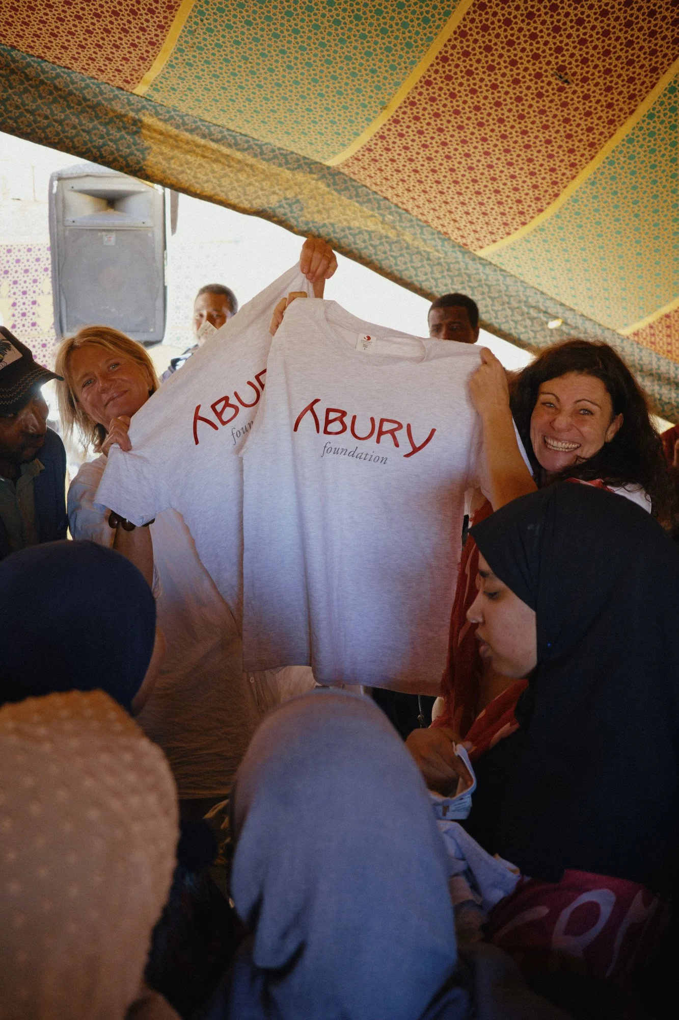 Menschen feiern eine Veranstaltung, halten T-Shirts mit dem Logo der Abury Foundation hoch, unter einem bunten Maisteppich.