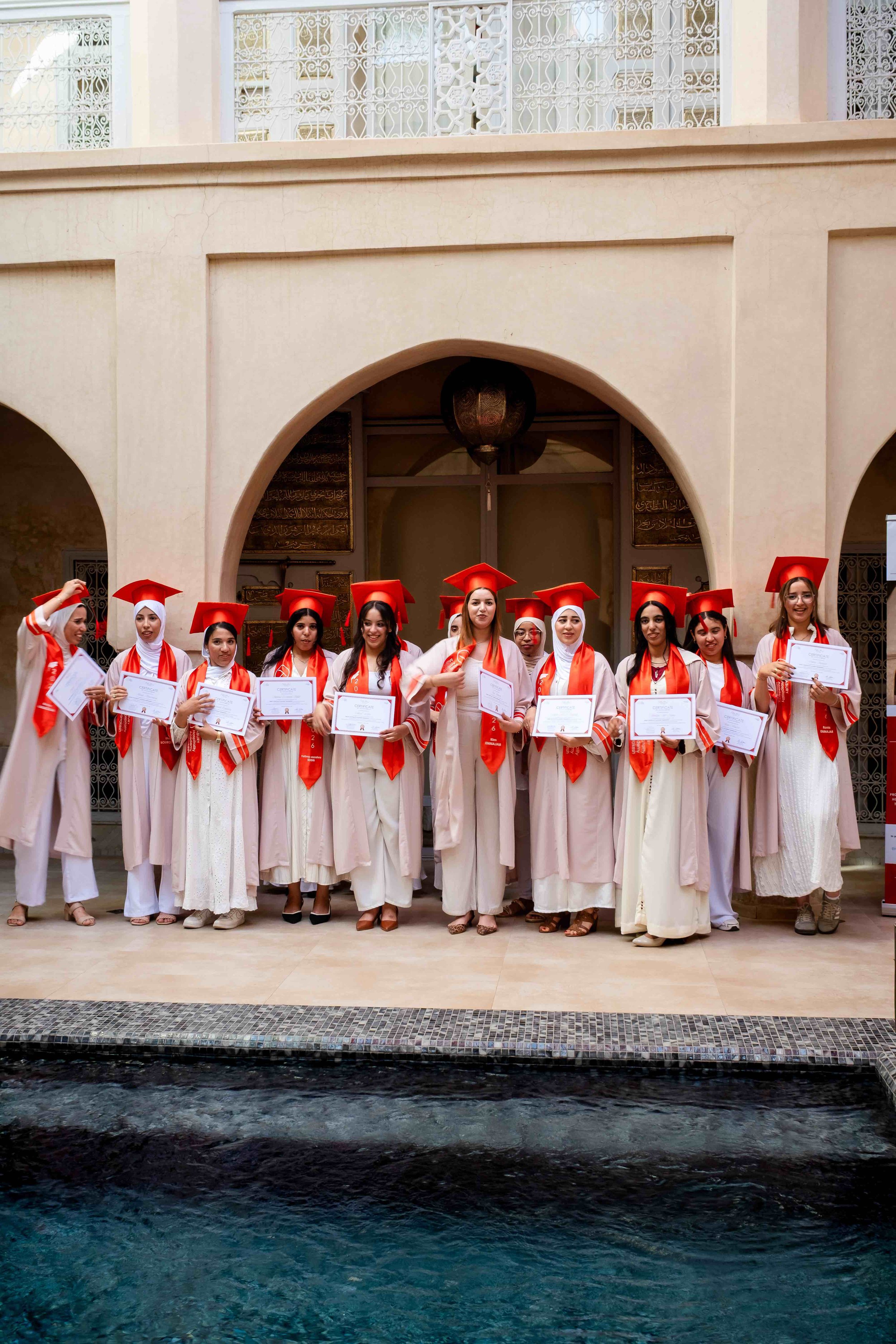 Gruppe von Absolventinnen in weißen Kleidern, roten Mützen und Schals, die bei einer Zeremonie ihre Abschlussurkunden erhalten haben, vor einem Gebäude mit arabischer Architektur und Ornamenten.