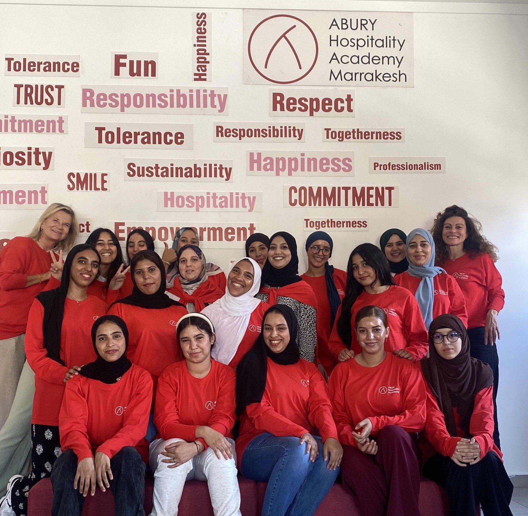 Gruppenfoto von Frauen und Mädchen, die rote T-Shirts tragen, vor einer Wand mit Wörtern wie Vertrauen, Verantwortung, Glück, Respekt, Nachhaltigkeit, Engagement und anderen. Das Bild wurde in einer Bildungseinrichtung aufgenommen.