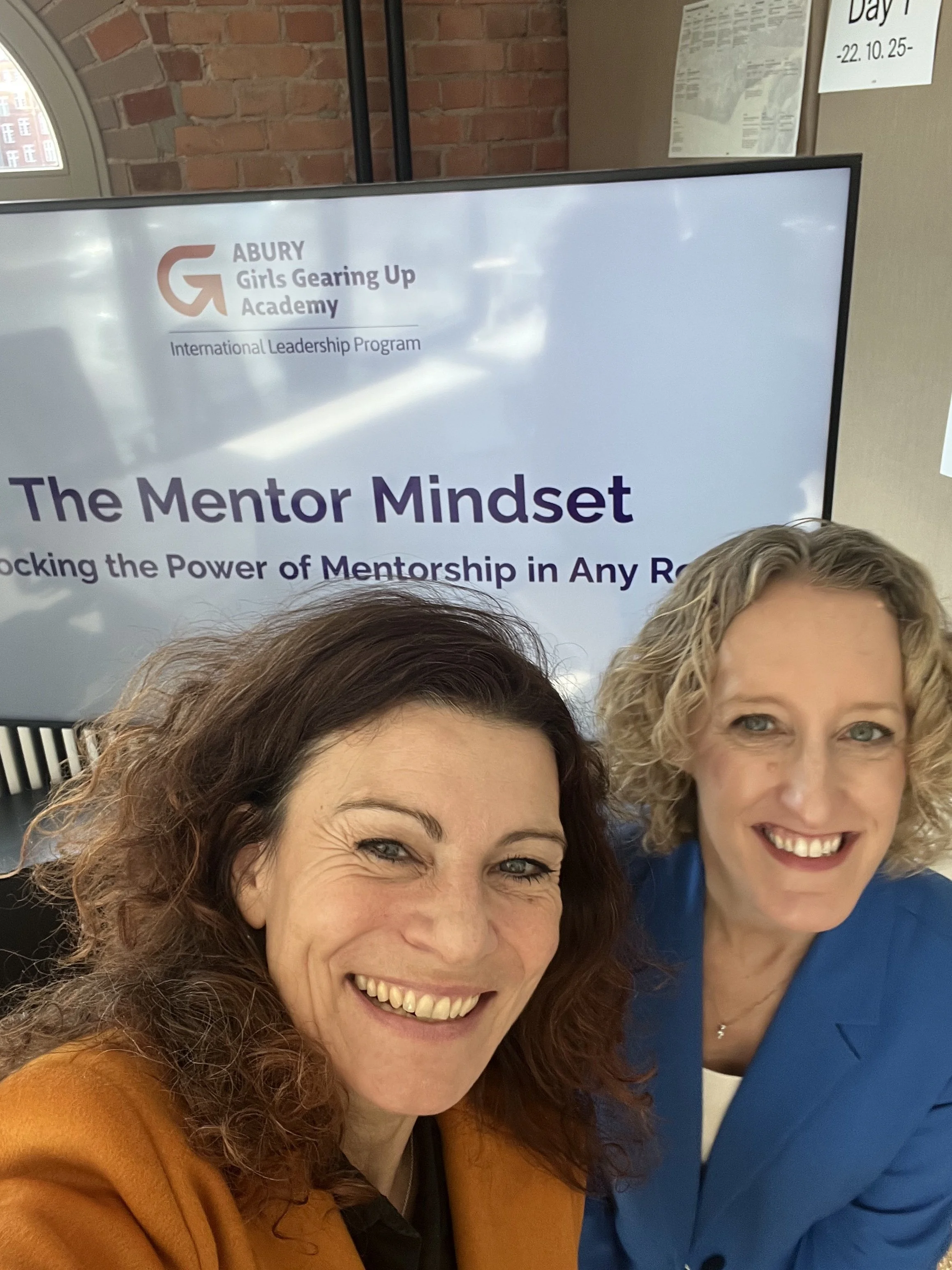 Zwei lachende Frauen vor einem Bildschirm, der das Logo von Girls Gearing Up Academy und den Titel „The Mentor Mindset“ zeigt.