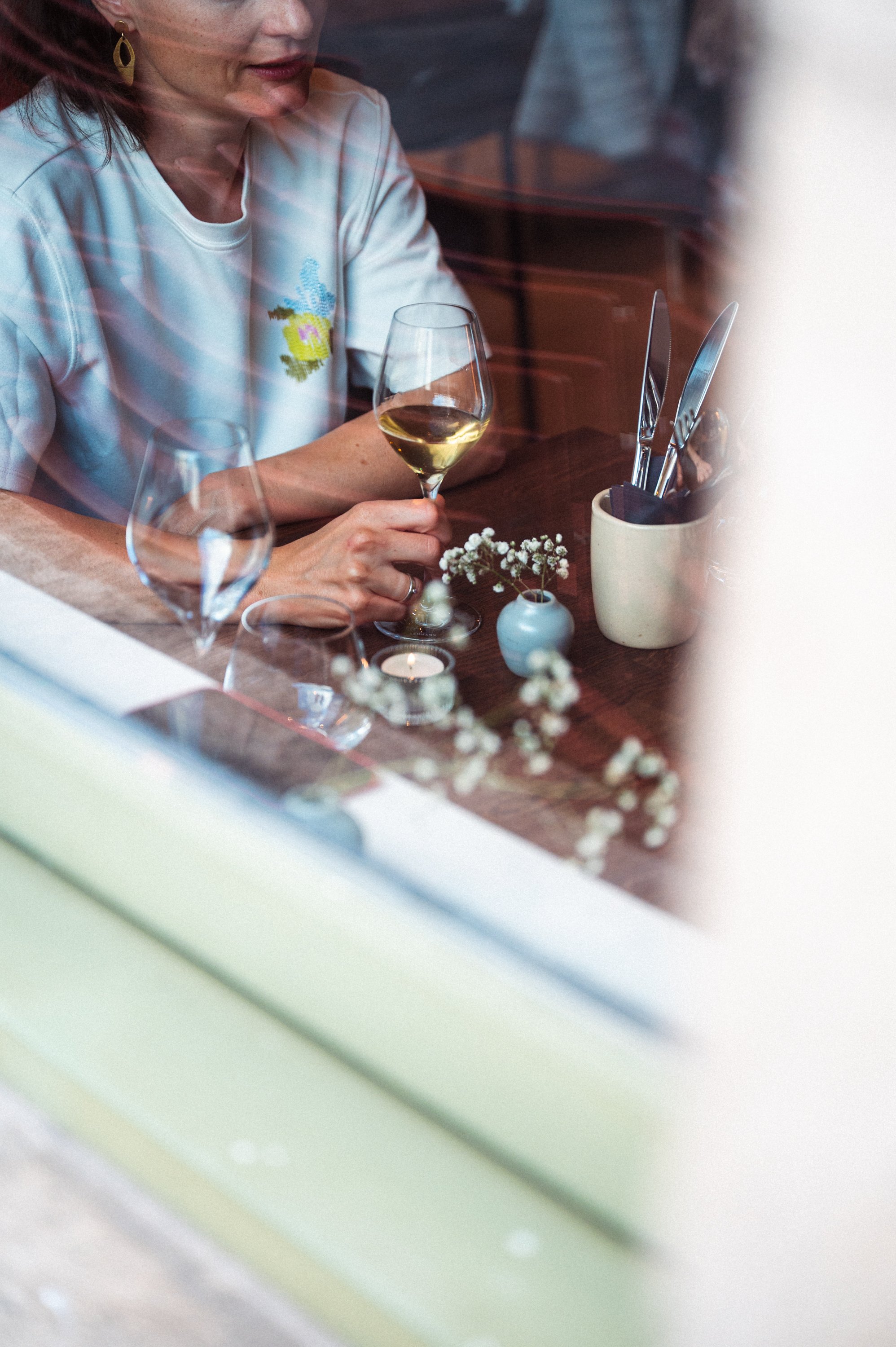 Een vrouw zit binnenachter een raam met een glas witte wijn in haar hand, op een tafel met wijn, bloemen en bestek.