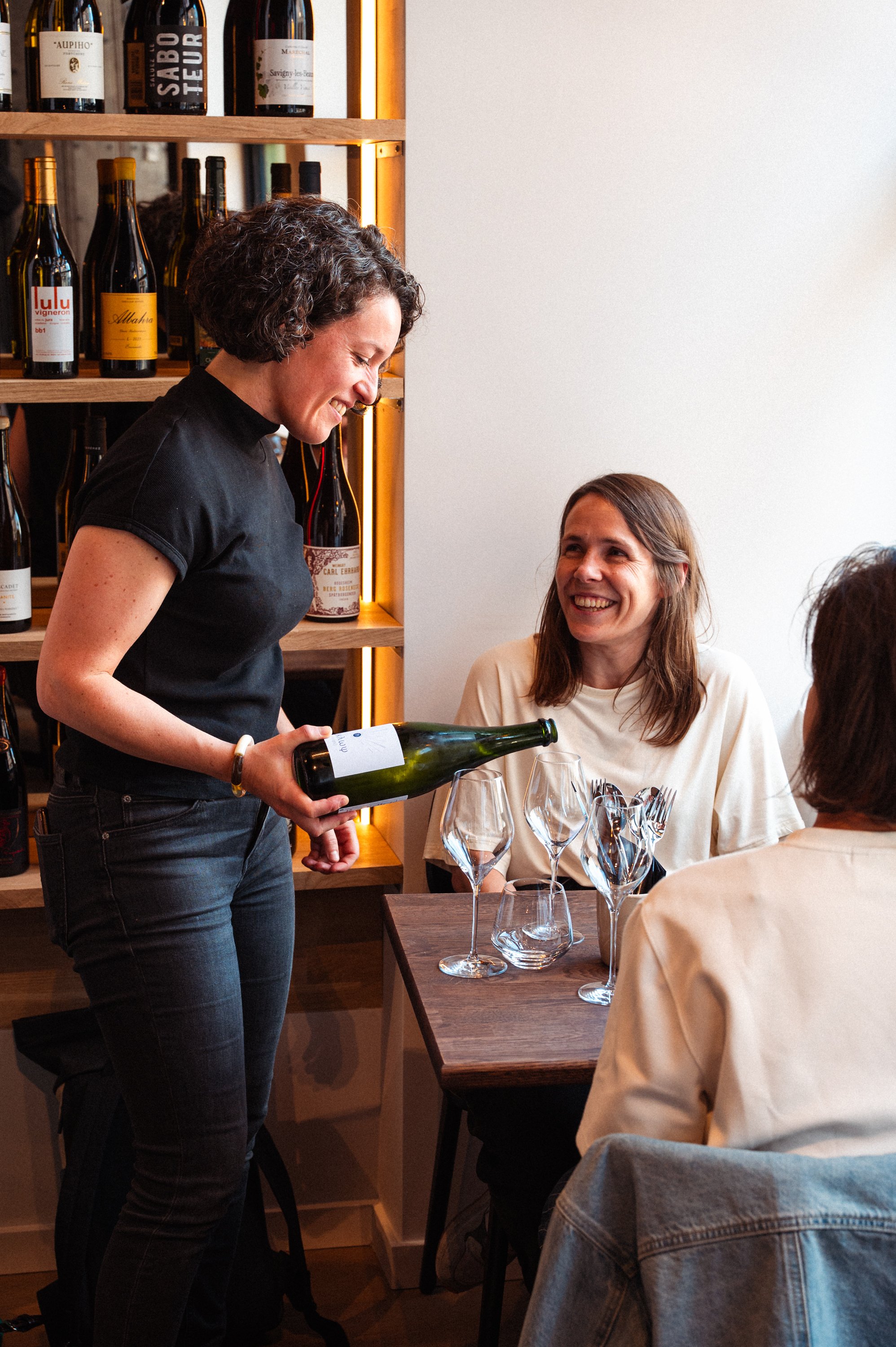 Vrouw schenkt wijn in glazen voor een tafel met drie mensen, lachende vrouw kijkt op terwijl ze wijn schenkt, wijnflessen op achtergrond.