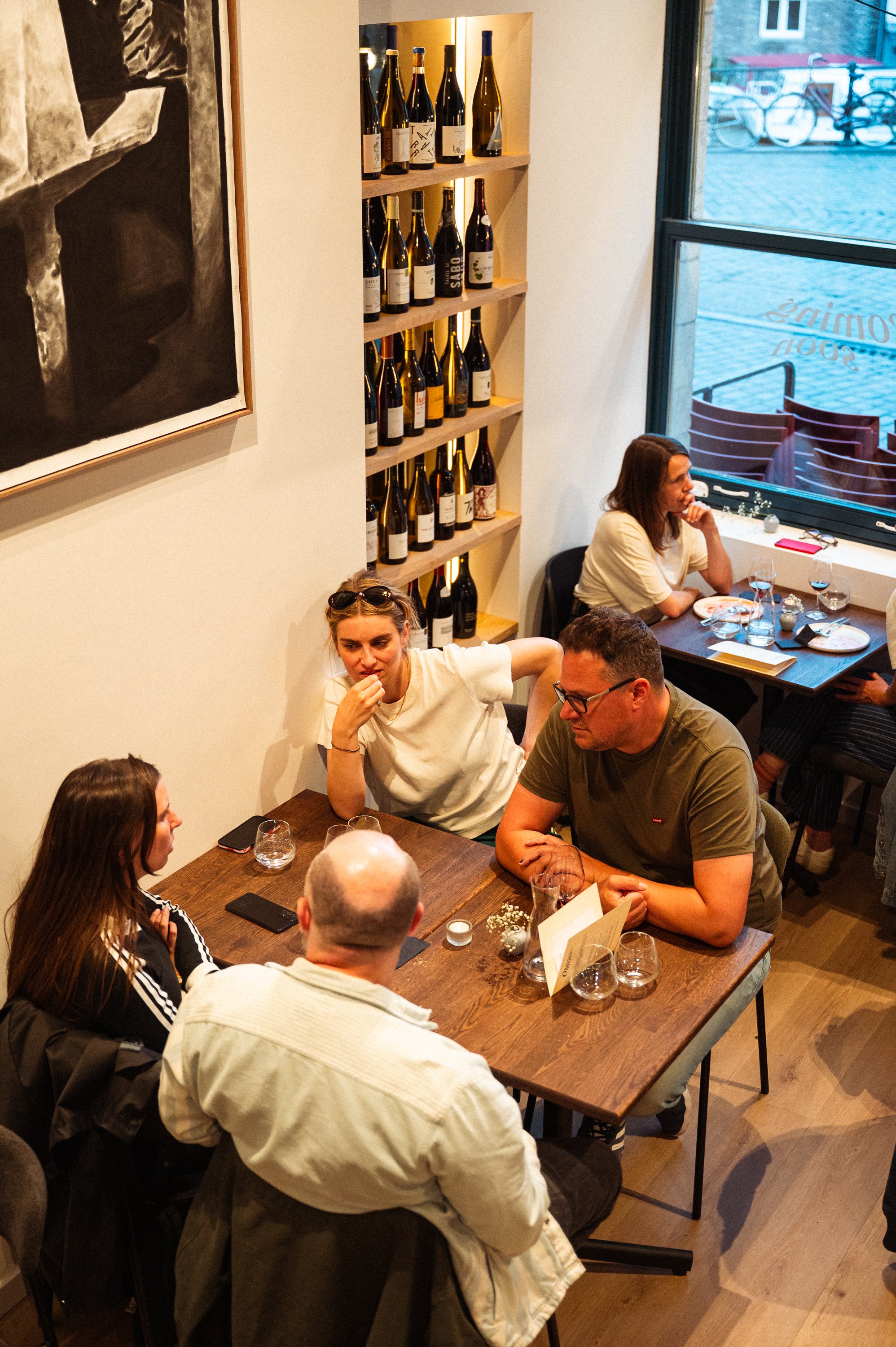 Binnen in een restaurant met een muur van wijnflessen en een grote afbeelding aan de muur, met meerdere groepen mensen aan tafels die eten en drinken.