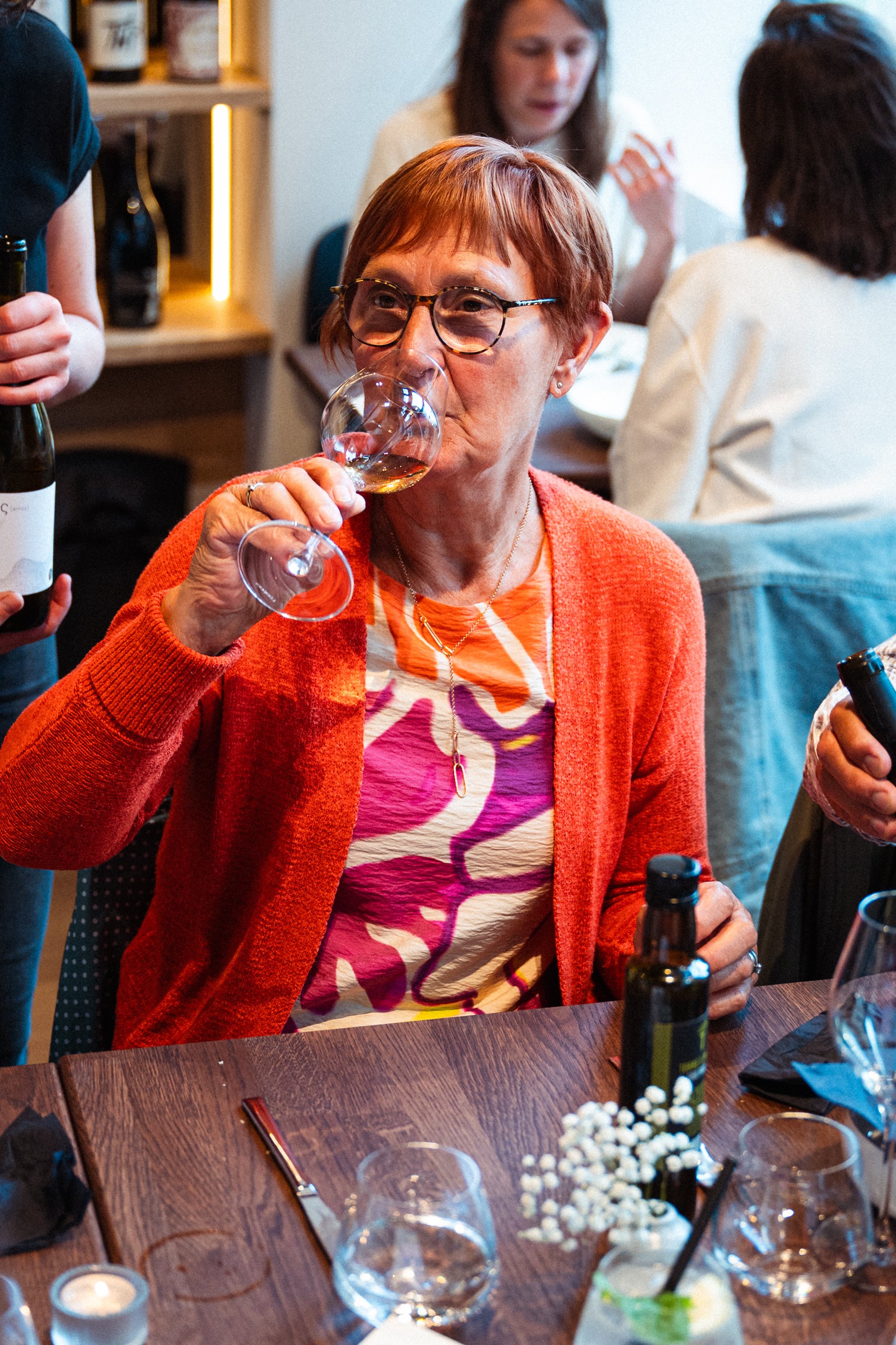 Vrouw drinkt witte wijn tijdens een etentje met vrienden of familie. Ze draagt een oranje cardigan en een kleurrijk shirt.