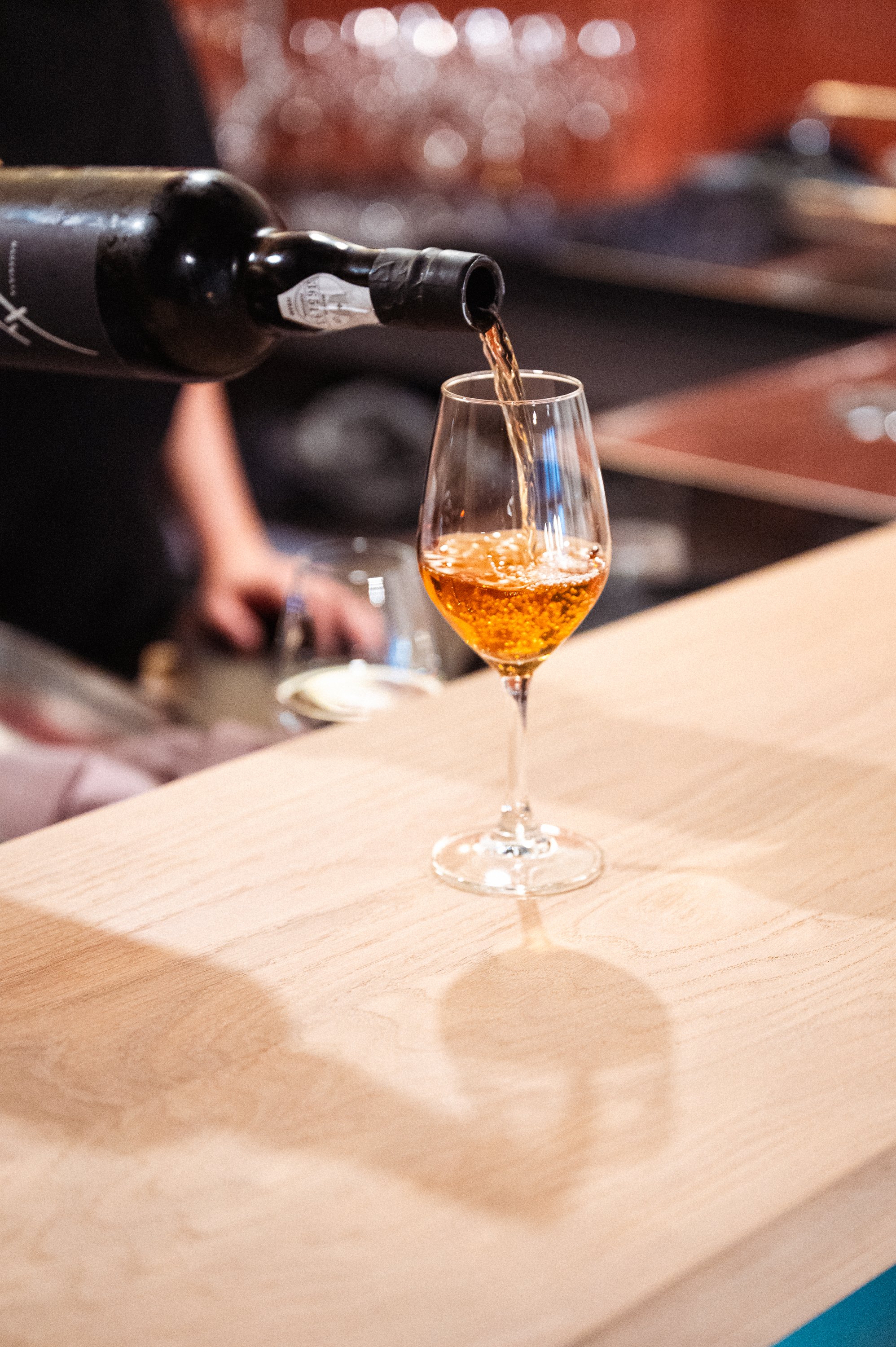 Een glas waarop rosé wijn wordt ingeschonken door een persoon, met een achtergrond van een bar of restaurant.