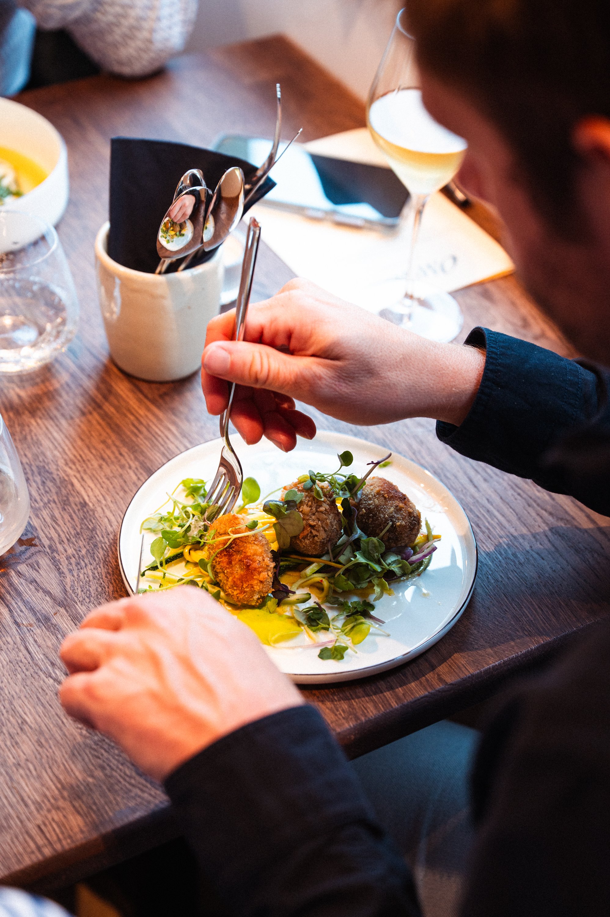 Persoon prikt kroketten op een bord met groene salade, op een houten tafel met wijn en water.