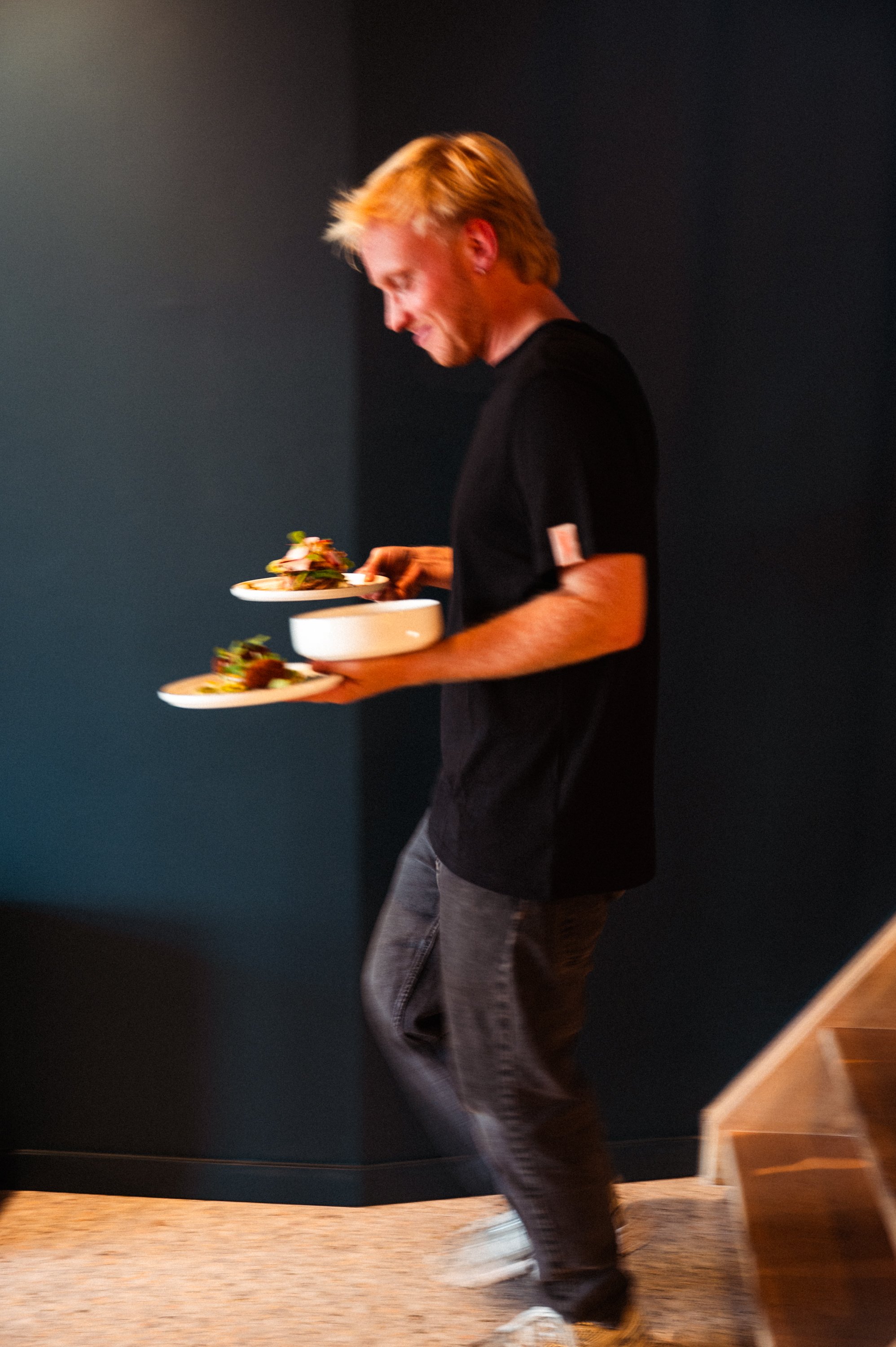 Man met blond haar, zwart shirt en spijkerbroek, loopt met twee borden vol eten langs een donker muur.