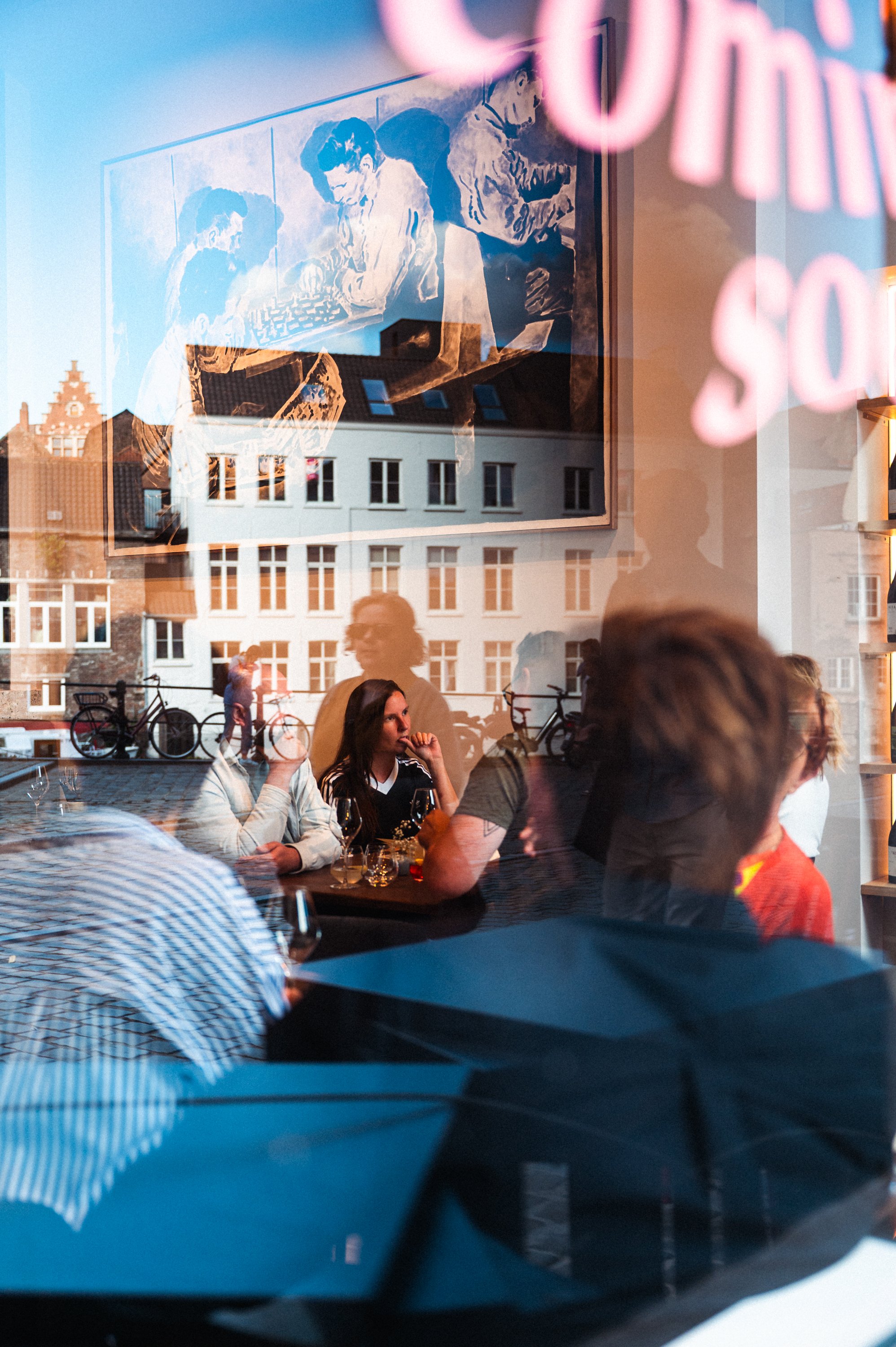 Reflexie van mensen en gebouwen in een etalaglas, met een binnen- en buitenzicht van een restaurant of café waar mensen zitten te praten en te drinken, buiten fietsen.