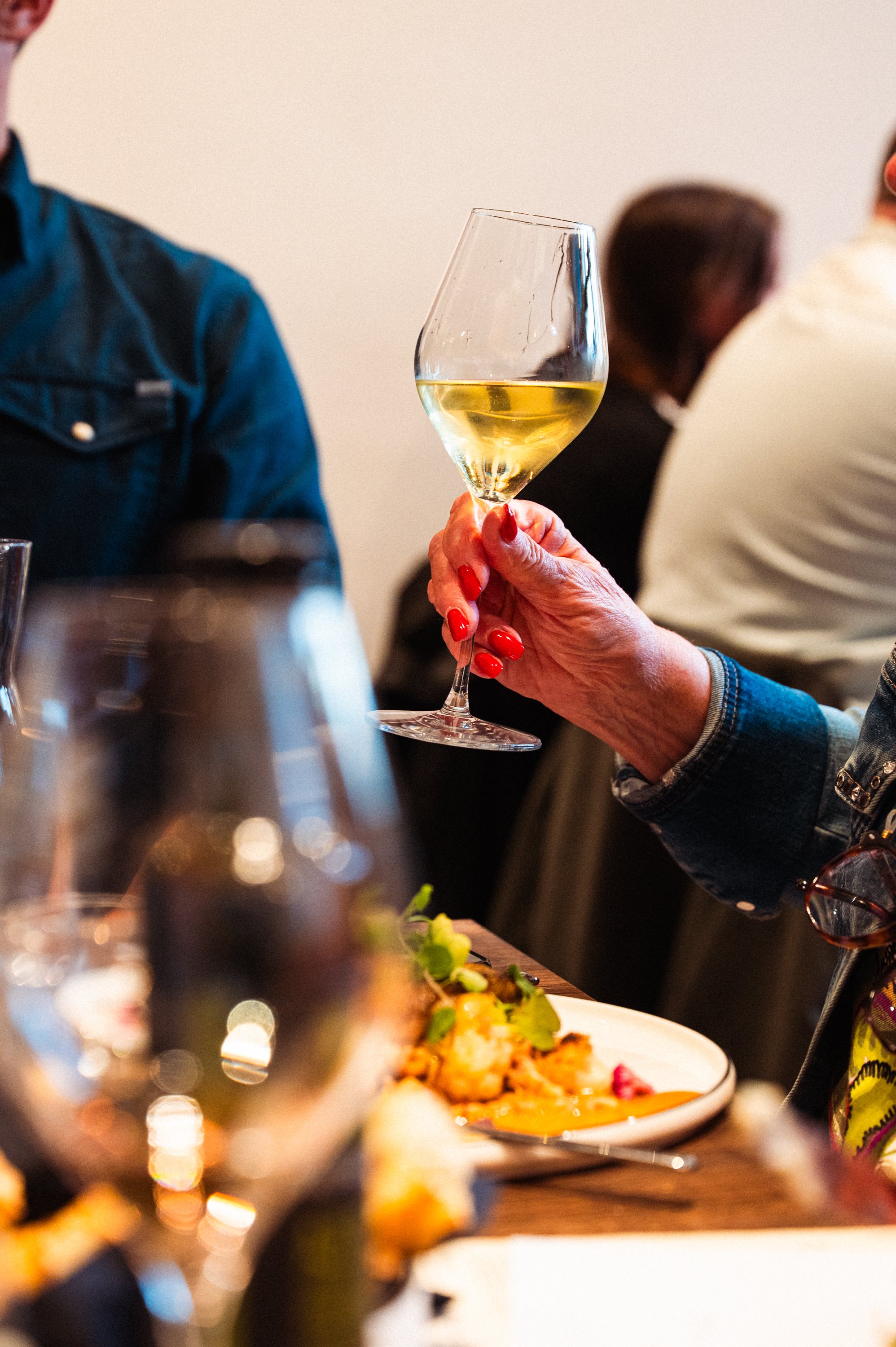 Een hand met rood gelakte nagels houdt een glas witte wijn vast tijdens een diner.