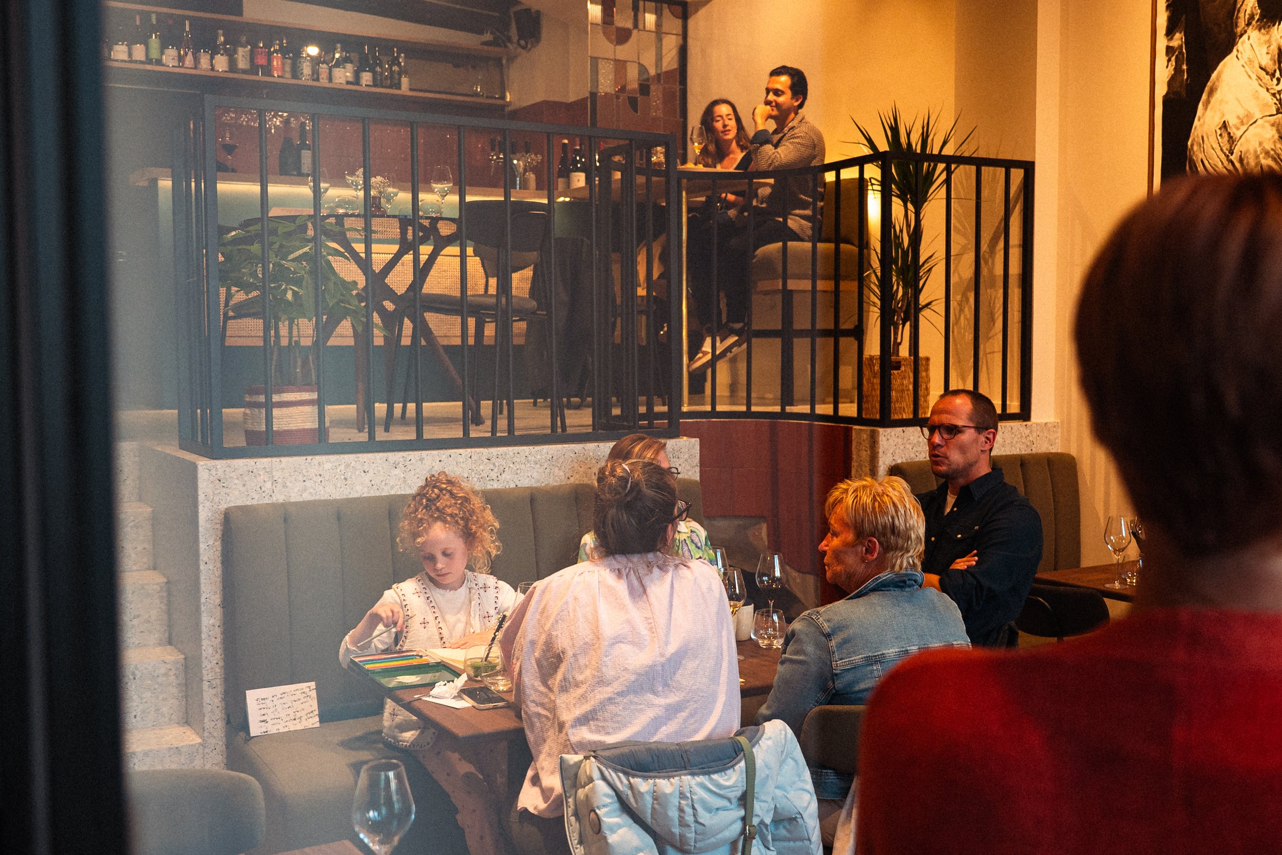 Een gezellig restaurant met mensen die eten en drinken. Op de tweede verdieping zitten twee mensen aan een tafel met glazen, terwijl op de onderste verdieping een groep van vijf mensen aan een tafel zitten, waaronder een kind die knutselt.