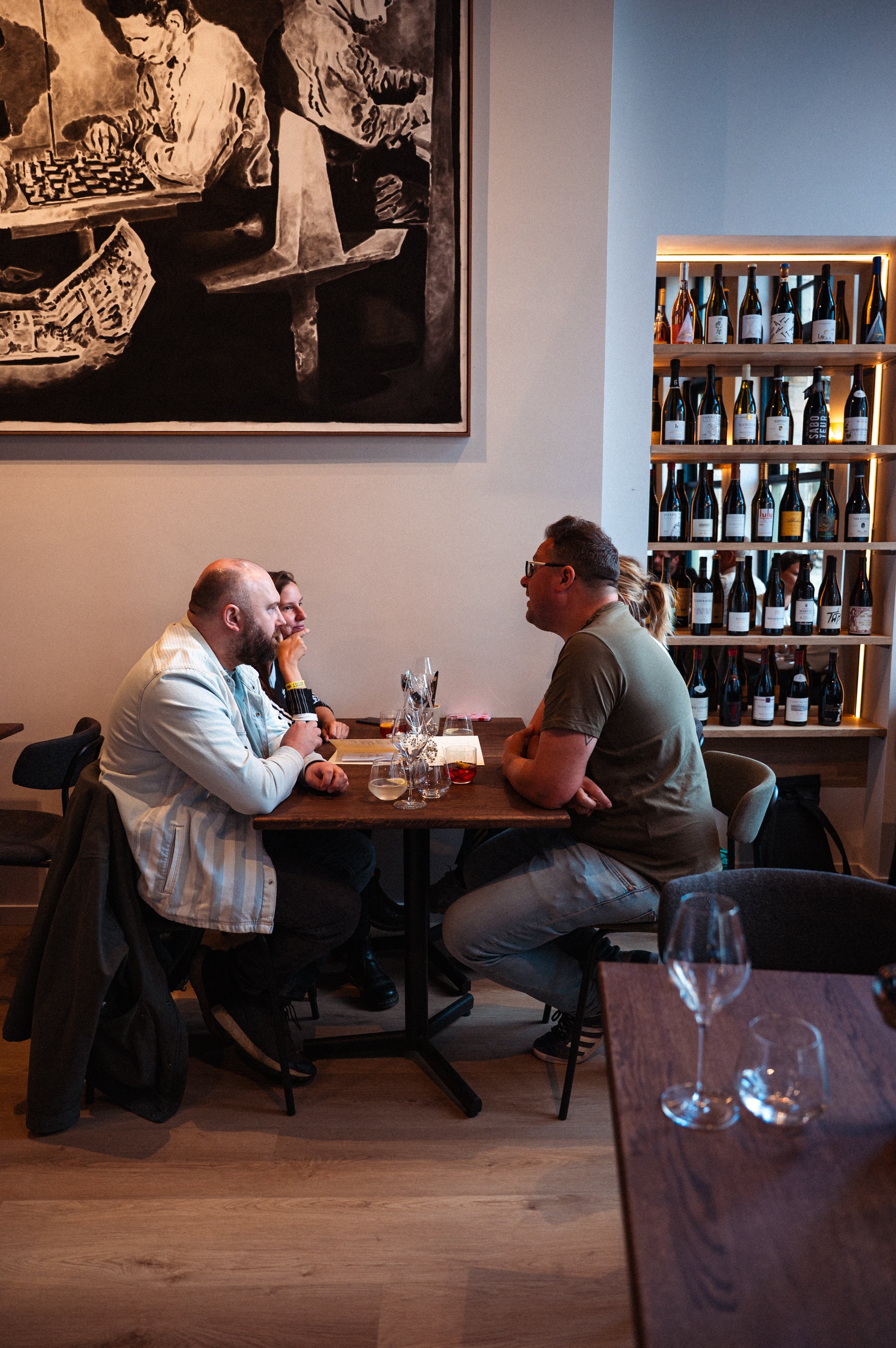 Vier mensen zitten rond een tafel in een wijnbar of restaurant, met wijnflessen op een houten wijnrek achter hen.