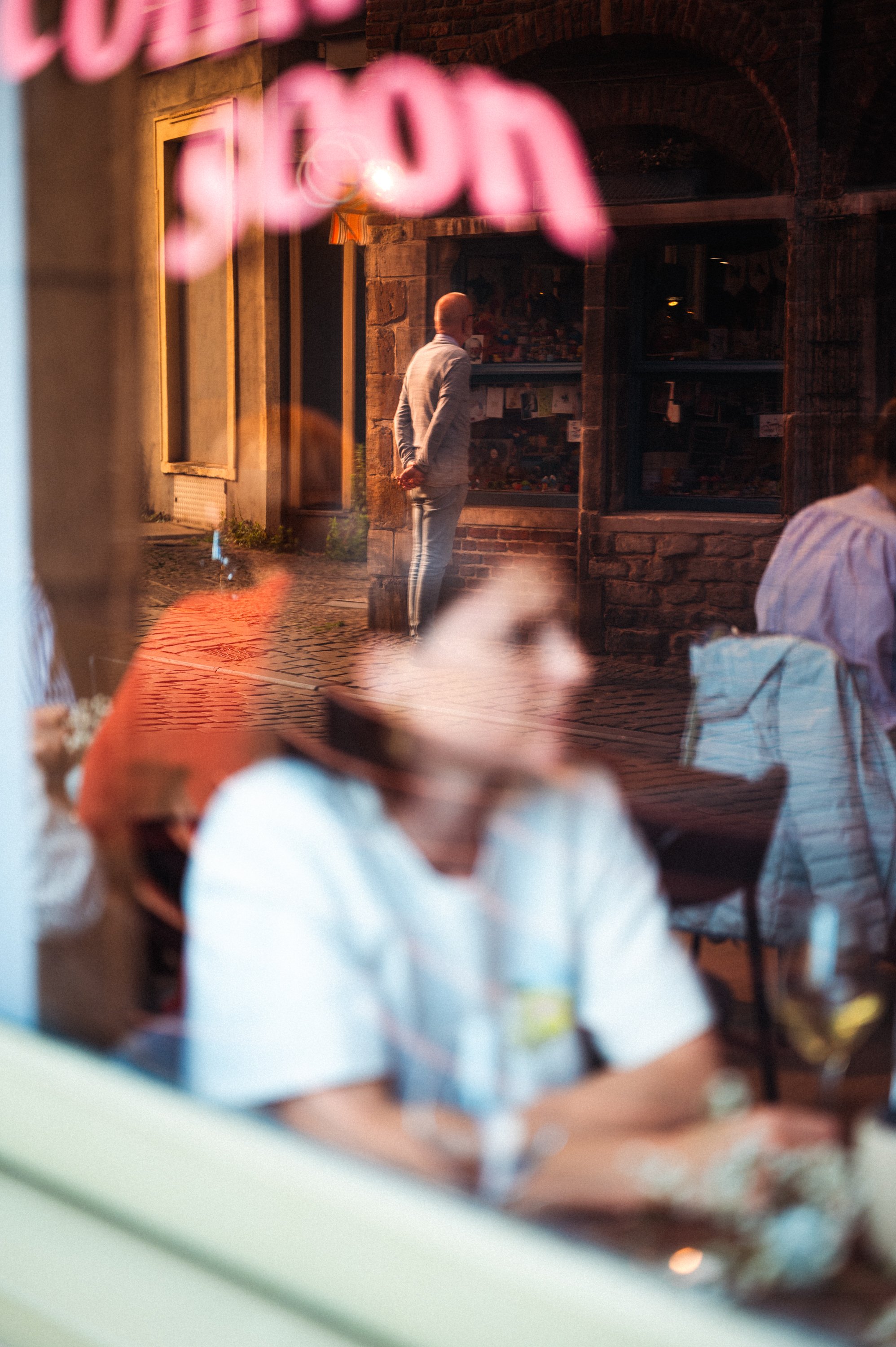 Foto van een straat met een persoon die naar een winkel kijkt, genomen door een raam dat reflecties van een restaurant op de voorgrond toont, met mensen aan een tafel in het restaurant.