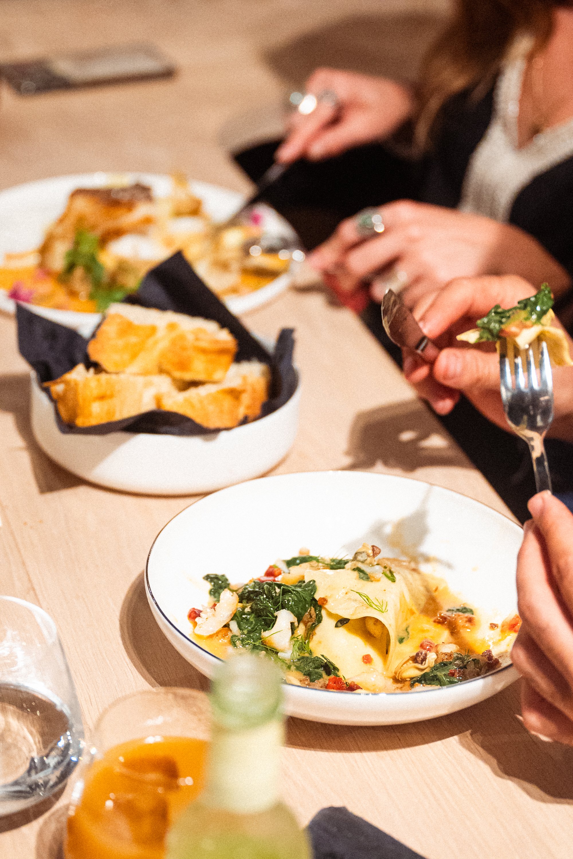 Mensen eten pasta met groenten in een restaurant. In het midden een wit bord met pasta en spinazie, en in de achtergrond een kom met knapperig stokbrood.