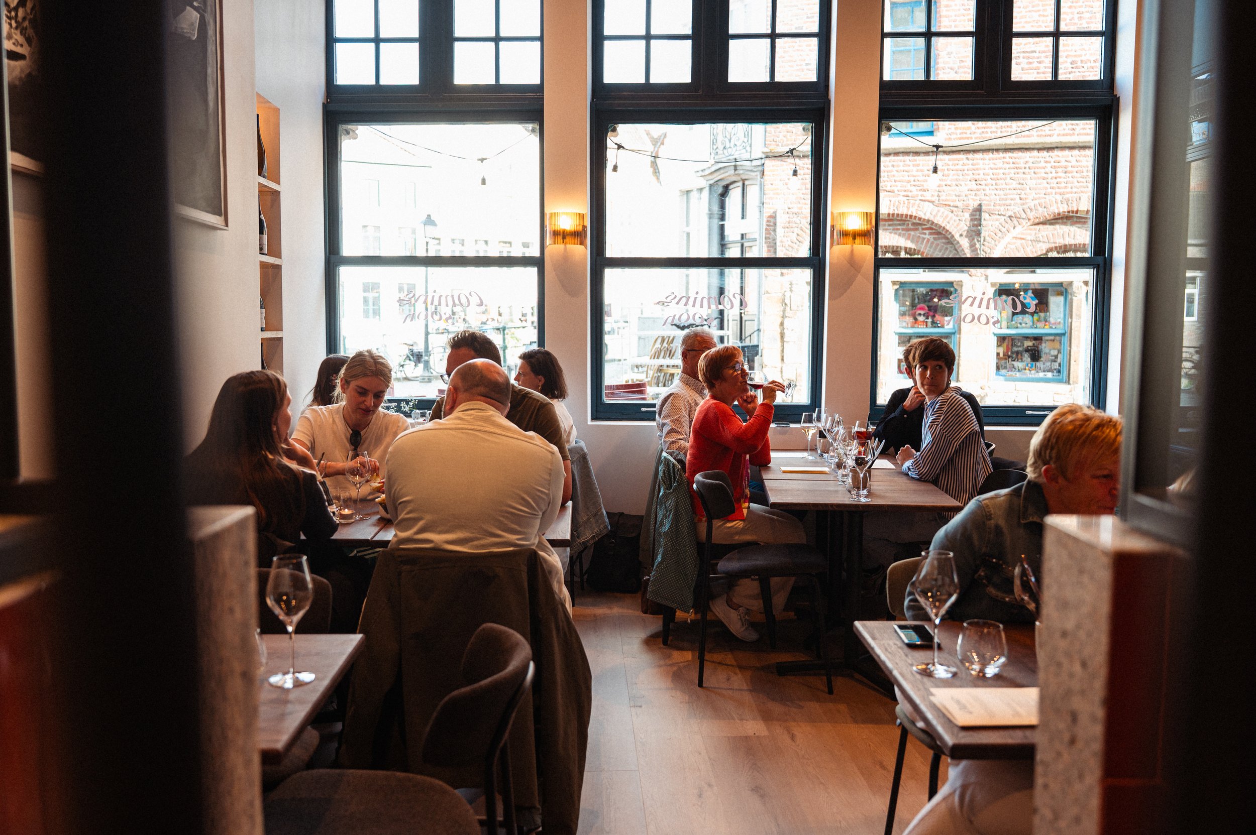Mensen zitten aan tafels in een restaurant met grote ramen en uitzicht op straat.