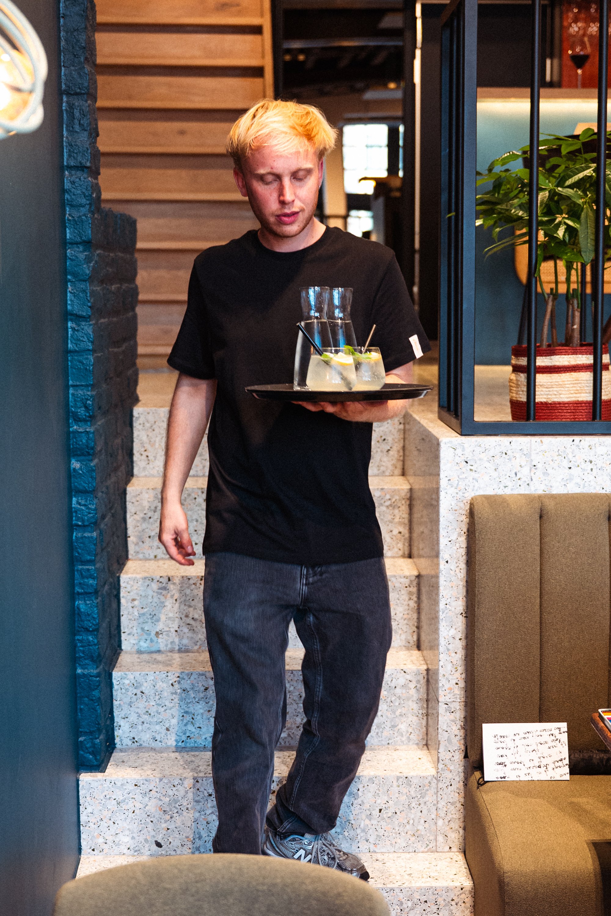 Een jonge ober met blond haar en lichte huid, gekleed in een zwart T-shirt en donkergrijze jeans, loopt de trap op in een restaurant of café, terwijl hij een zwarte dienblad met twee glazen limonade met citroen en munt vasthoudt.