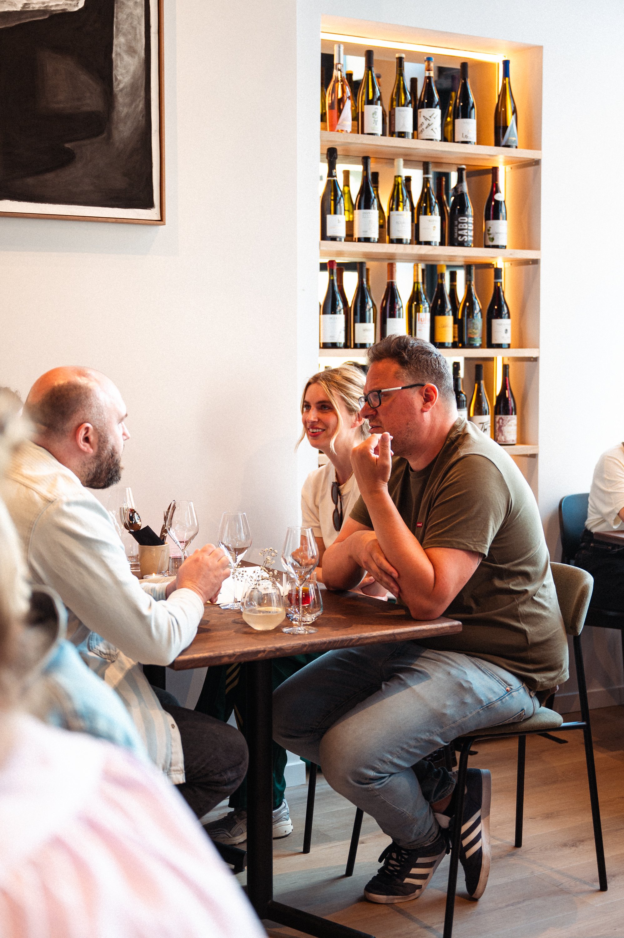 Drie mensen zitten aan een tafel in een restaurant, wijnflessen op een houten plank aan de muur, wijn- en drinkglazen op tafel, mensen praten en lachen.