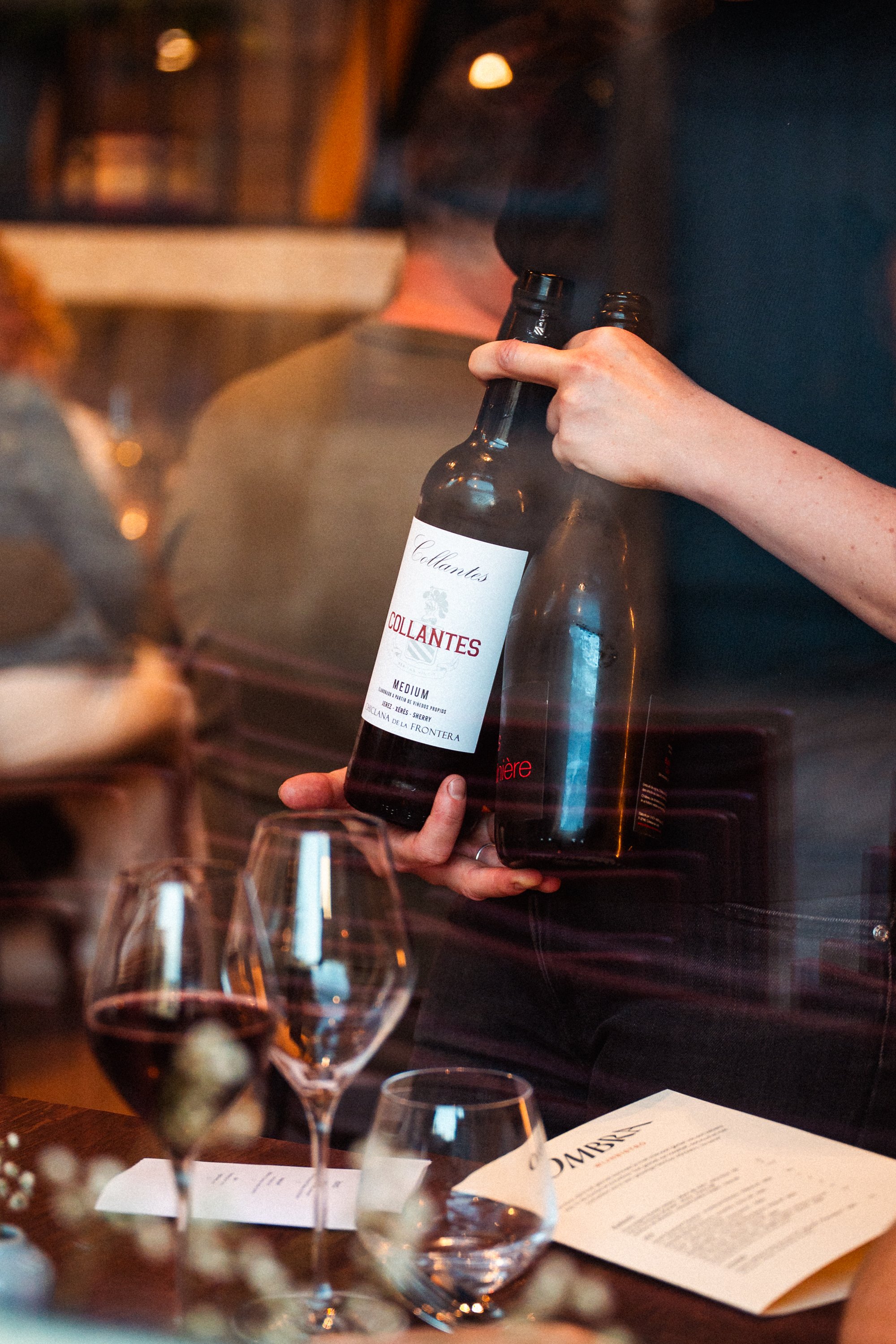 Persoon houdt twee flessen rode wijn vast met een glas wijn en een menu op tafel, binnen een restaurant of wijnbar.