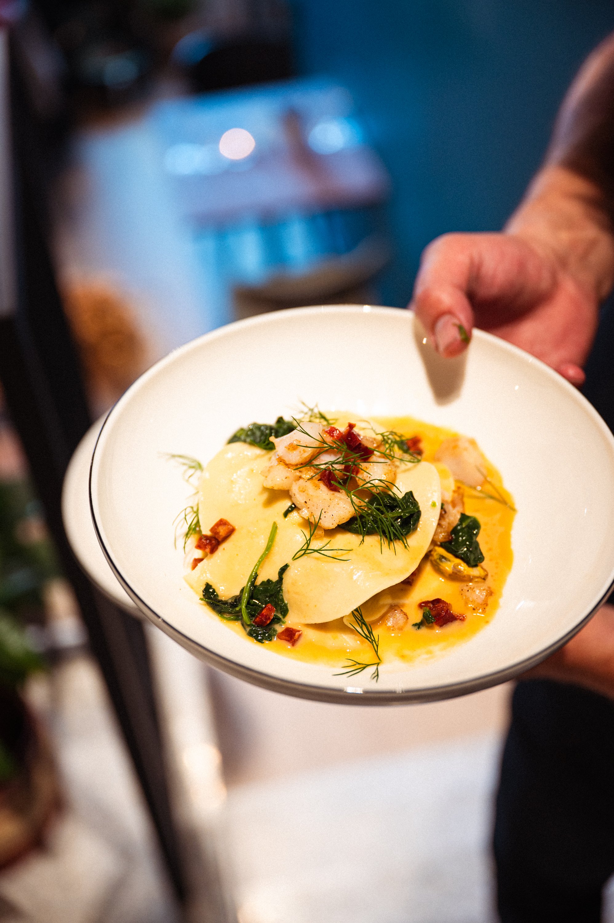 Close-up van een bord met ravioli, gekookt met groene groenten en saus, garnering met kruiden en stukjes bacon, door iemand aangeboden.