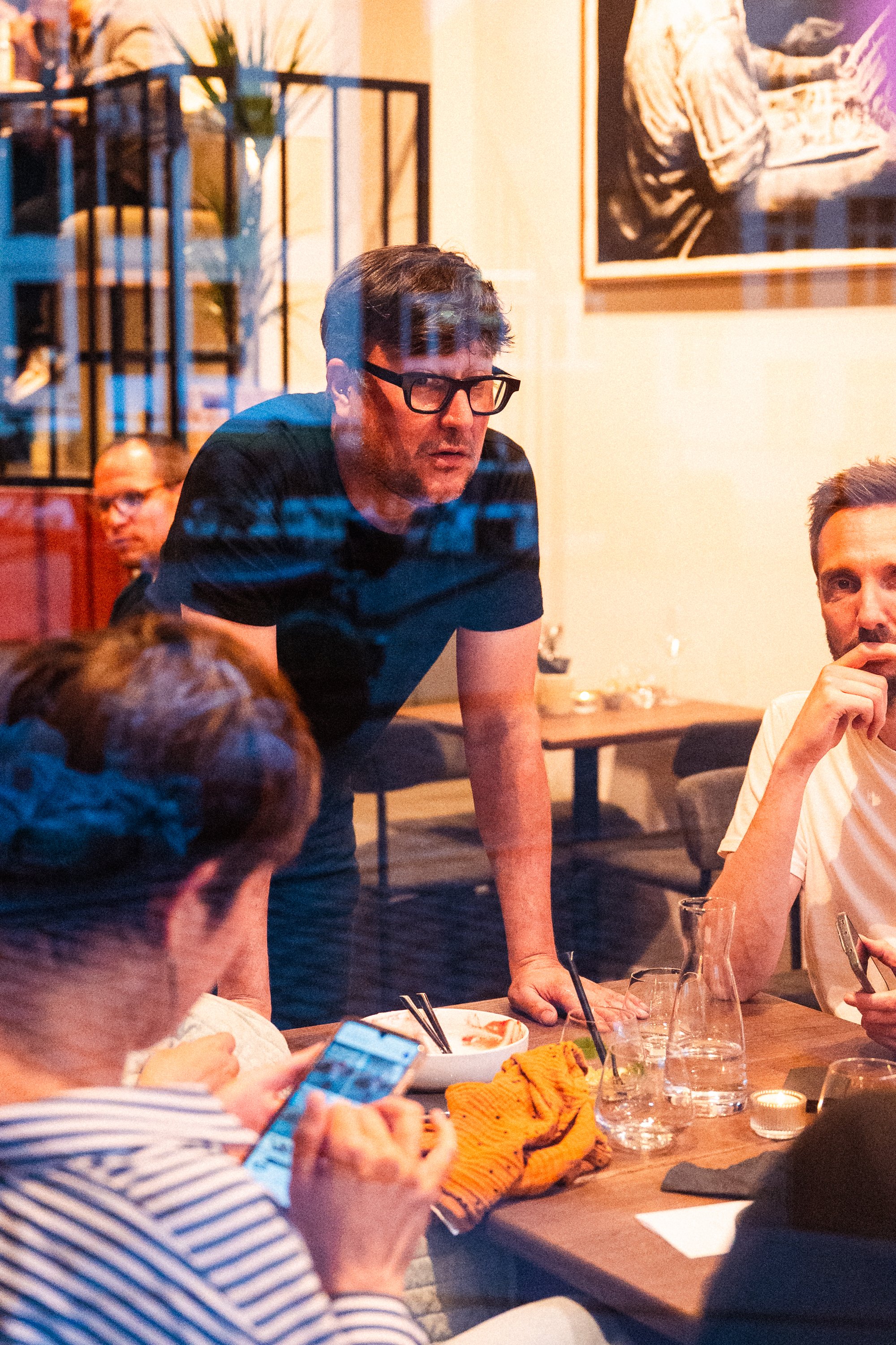 Mannen met zwarte haar en bril die zich bezighoudt met een gesprek aan een eettafel in een restaurant, omringd door andere mensen.