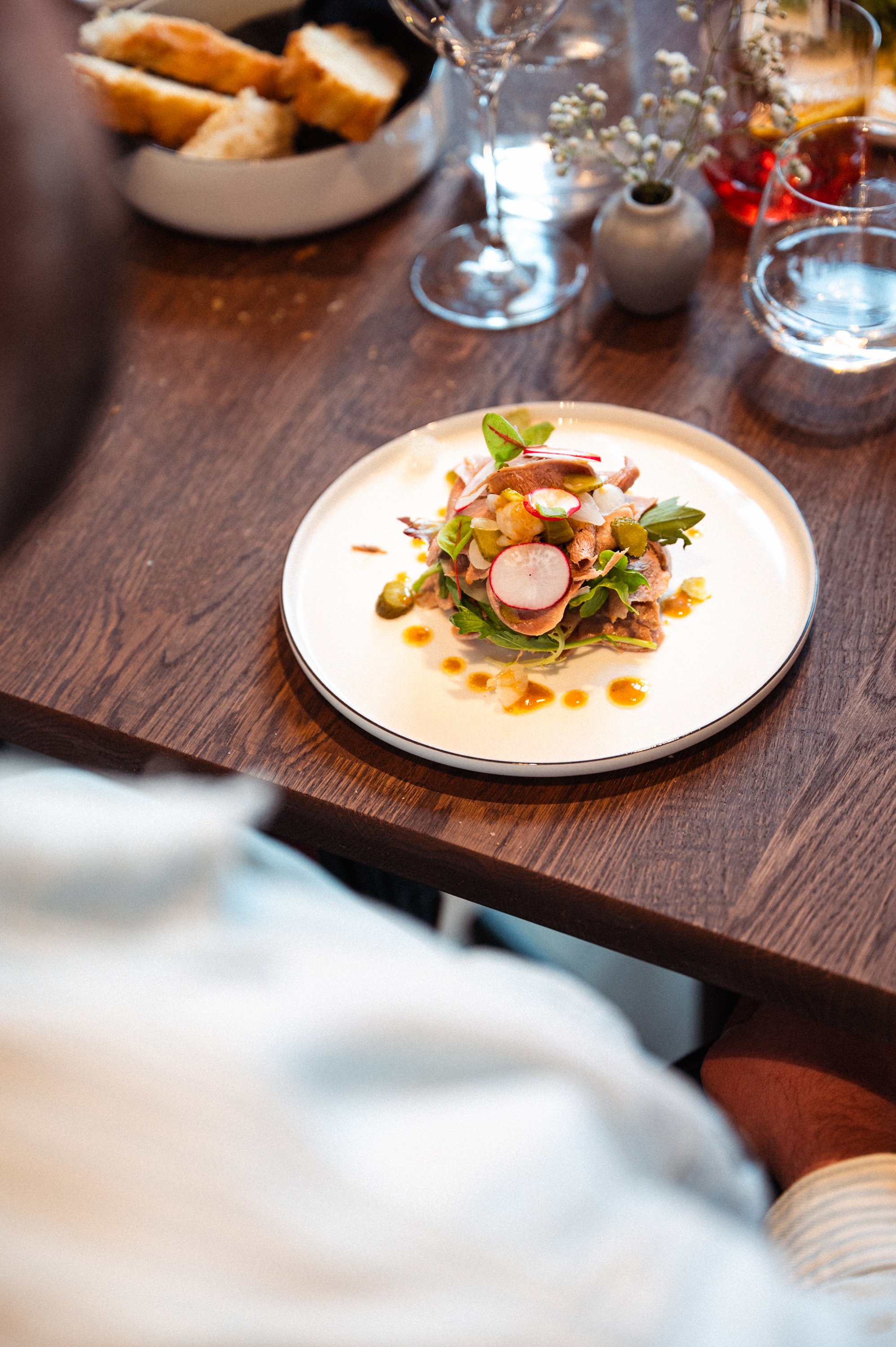 Een bord met een salade met radijs, dun gesneden vlees en groene bladgroenten, versierd met een dressing. Op de achtergrond een kom met brood, glazen en kleine vazen met bloemen op een houten tafel.