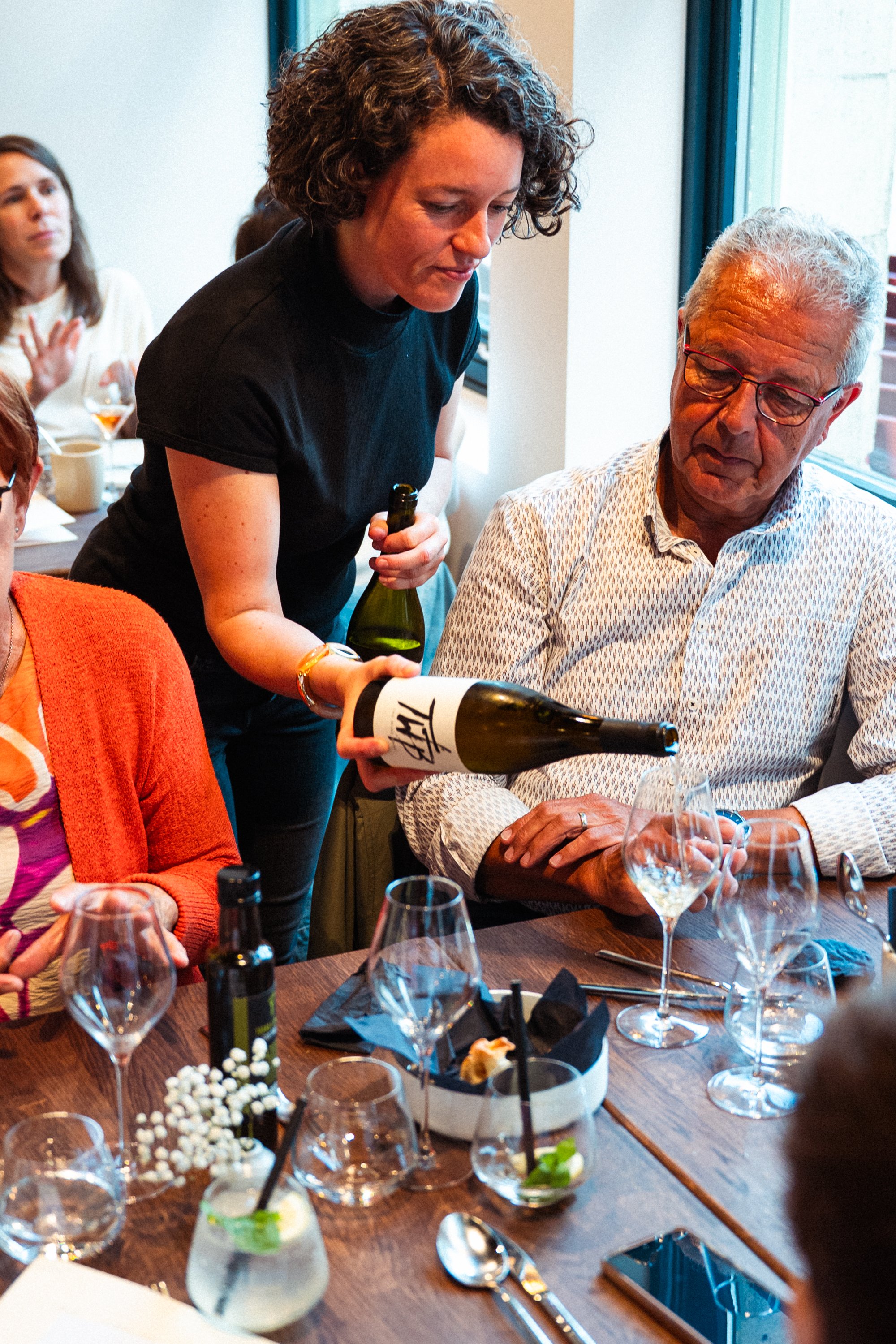Vrouw schenkt wijn in een glas tijdens een etentje met meerdere mensen aan een houten tafel.