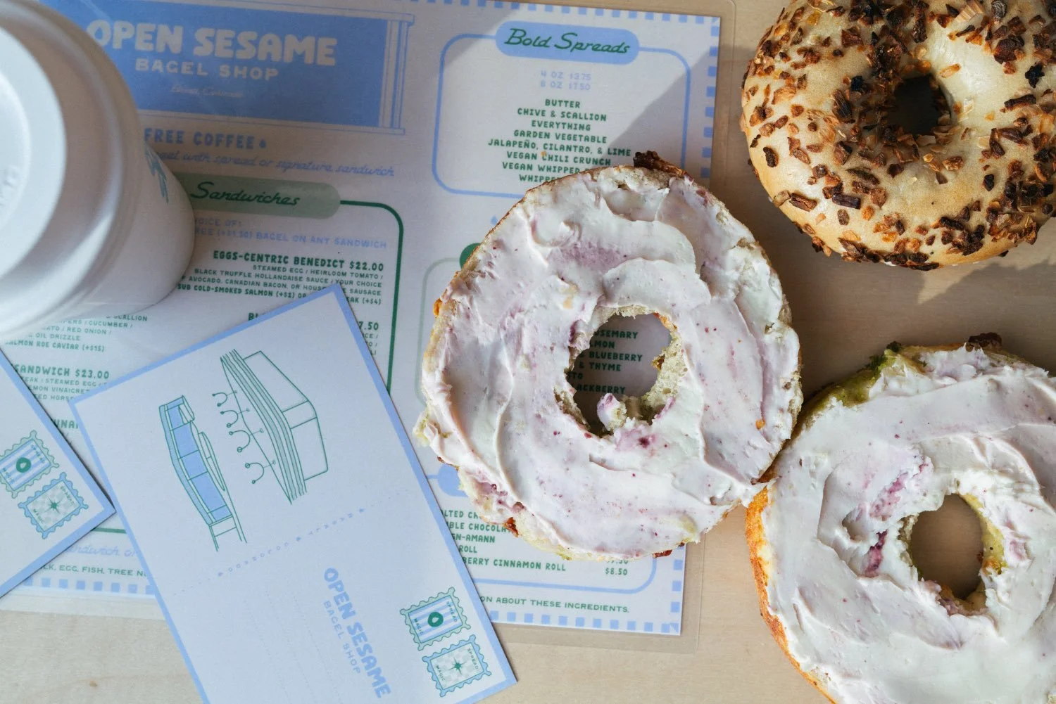 Two glazed donuts with frosting, one sprinkled with chocolate chips and the other with a different frosting, placed on a wooden surface alongside a menu, a coffee cup, and a business card from Open Sesame Bagel Shop.