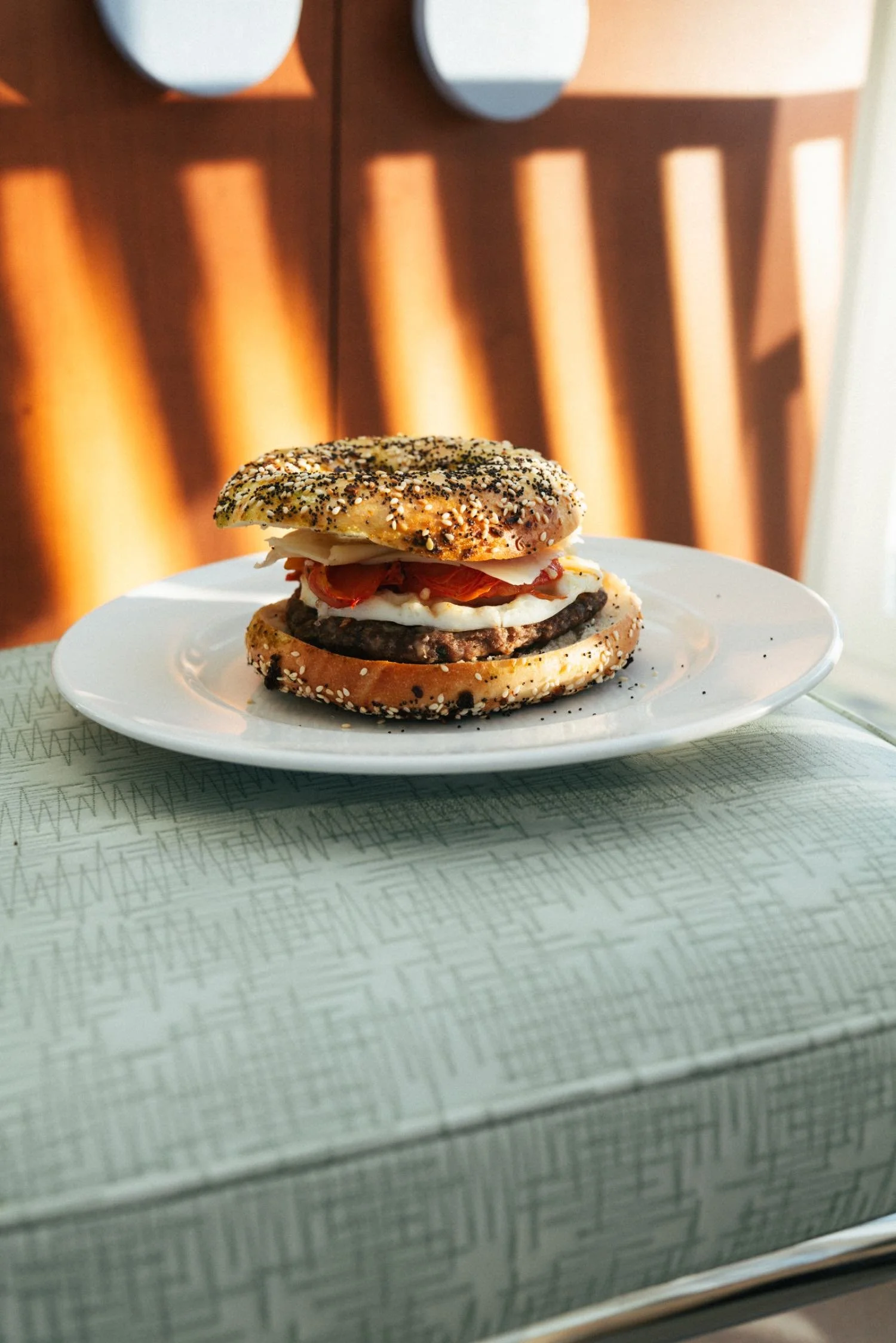 A cheeseburger on a white plate, with sesame seed bun, cheese, tomato, lettuce, and a beef patty, inside a restaurant with sunlit window blinds.