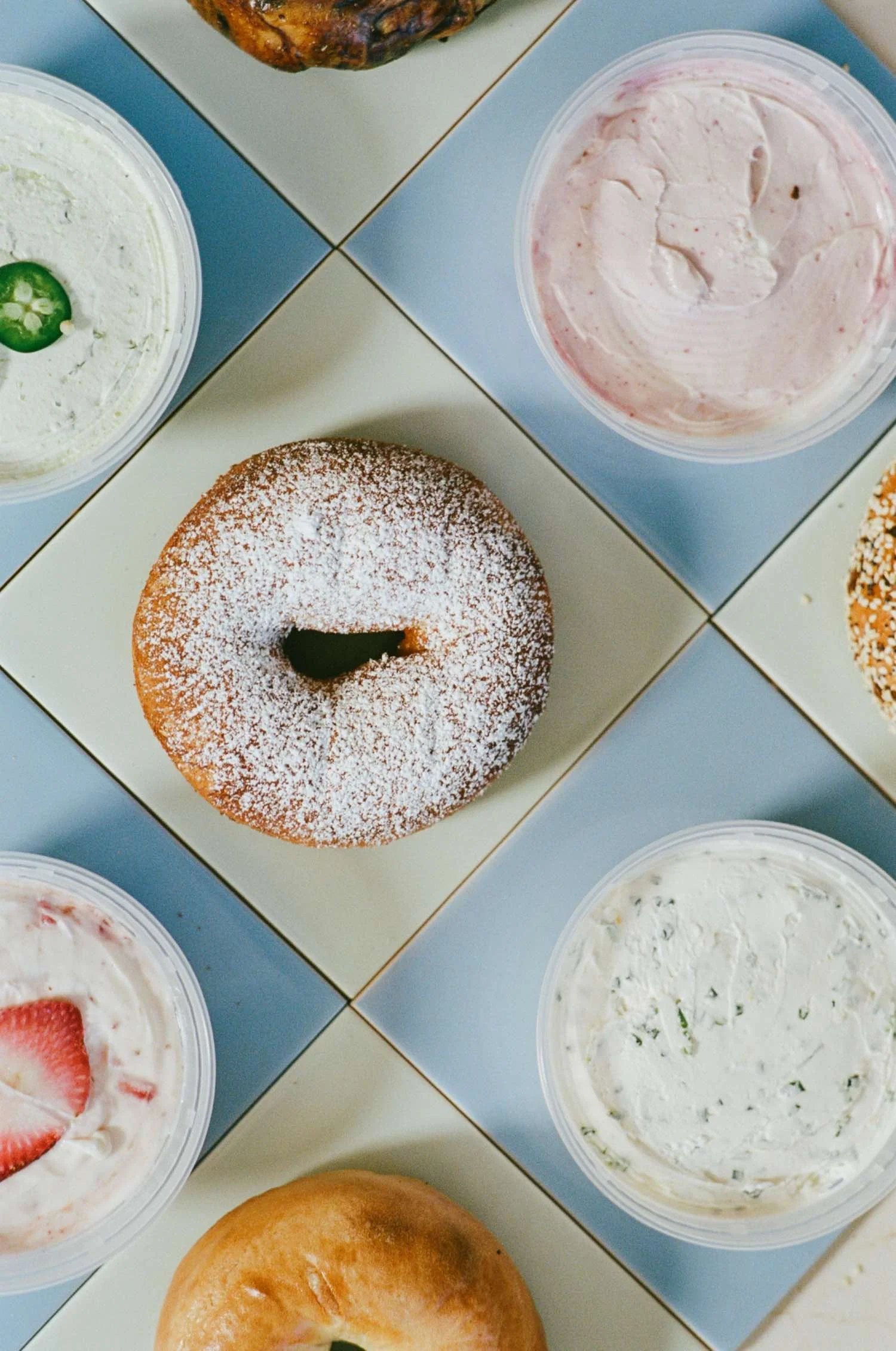 Multiple bowls of creamy dips, some garnished with strawberries, jalapeños, and herbs, along with a glazed donut and a toasted bread roll on a tiled surface.