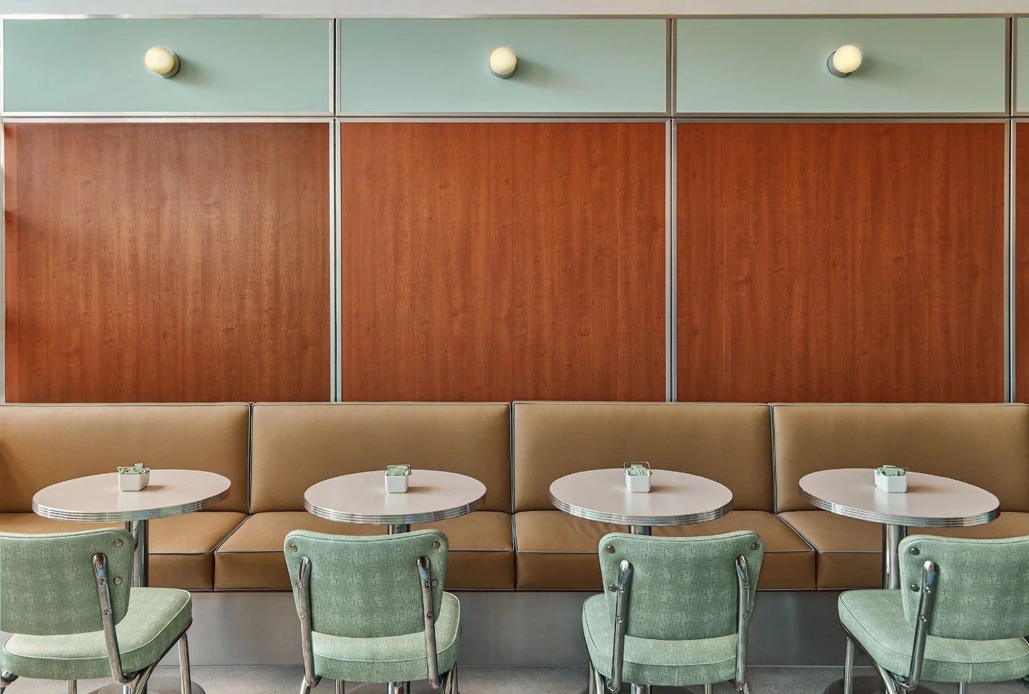 Empty vintage-style diner booth with three round tables and four green chairs, beige cushioned bench, and wooden wall paneling with three small lights.