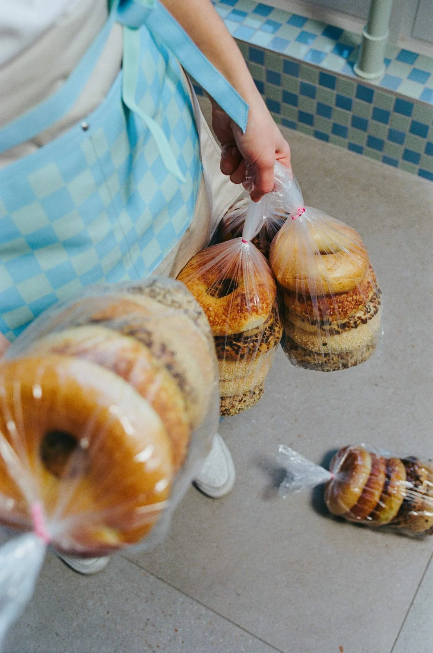 A person holding several bags of assorted pastries, including donuts and cinnamon rolls, in a bakery or cafe setting with tiled walls and a gray floor.
