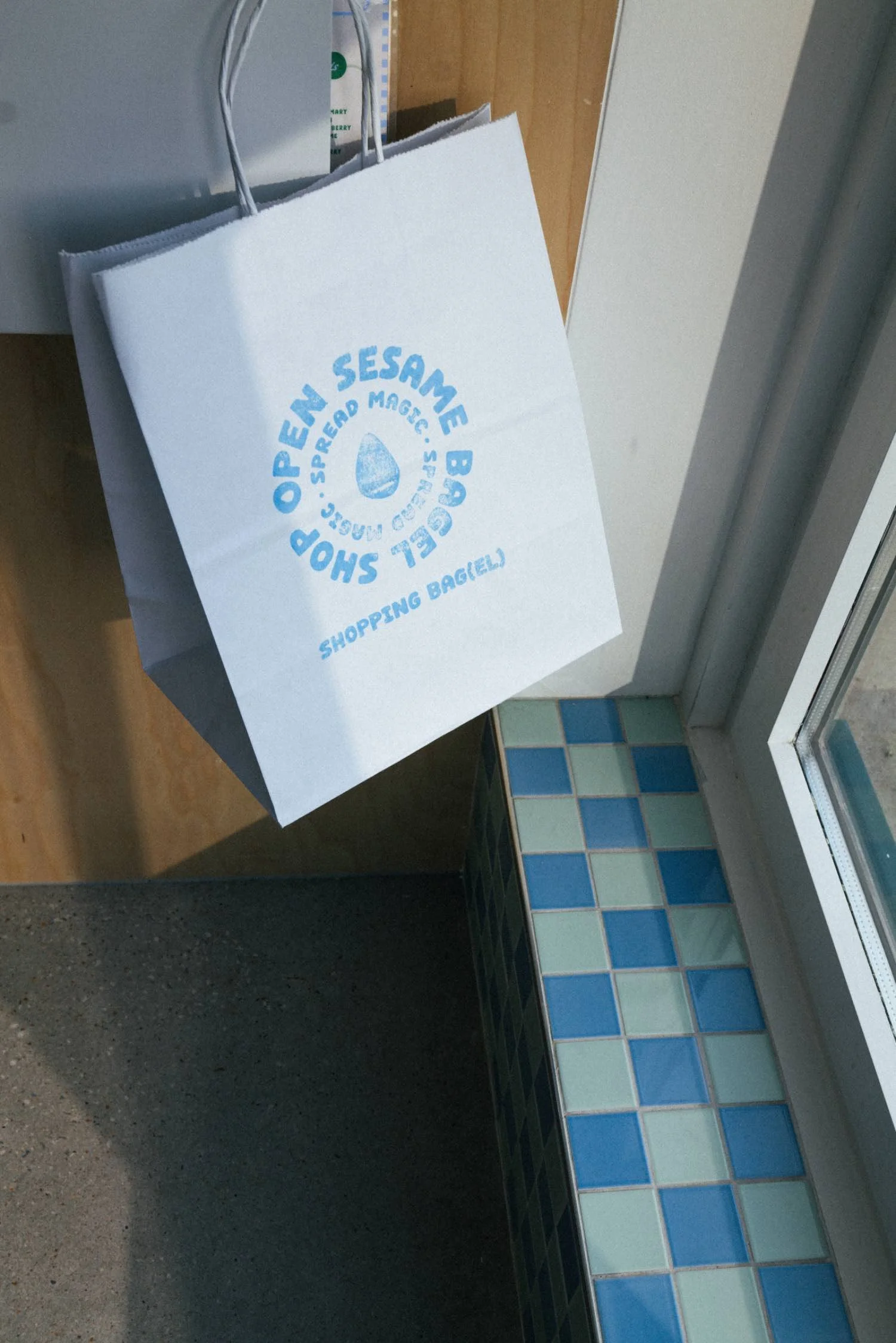 White shopping bag with blue logo hanging from a hook near a tiled wall and glass window, on a countertop.