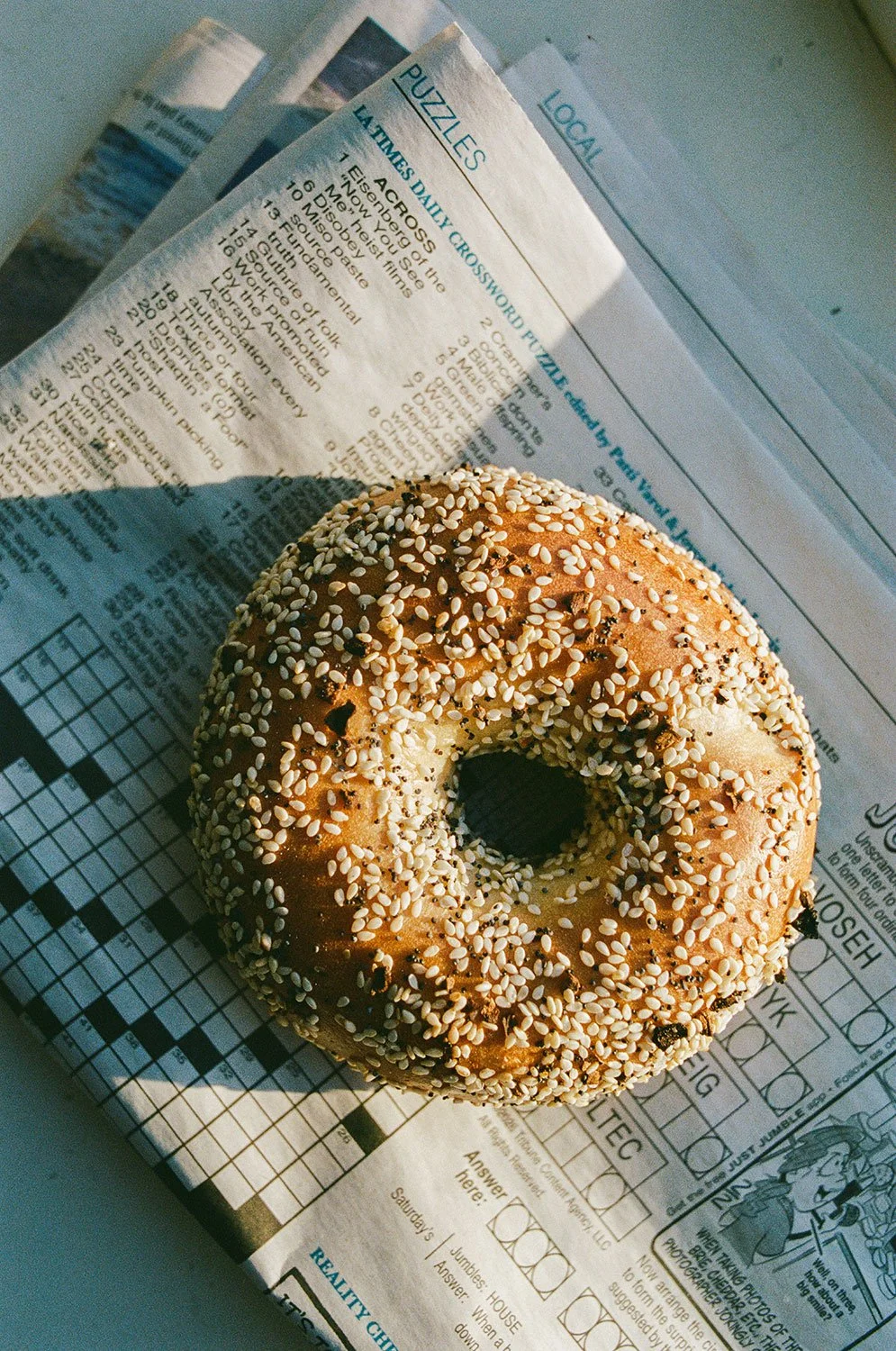 A sesame seed bagel on a tray of newspaper with crossword puzzles.